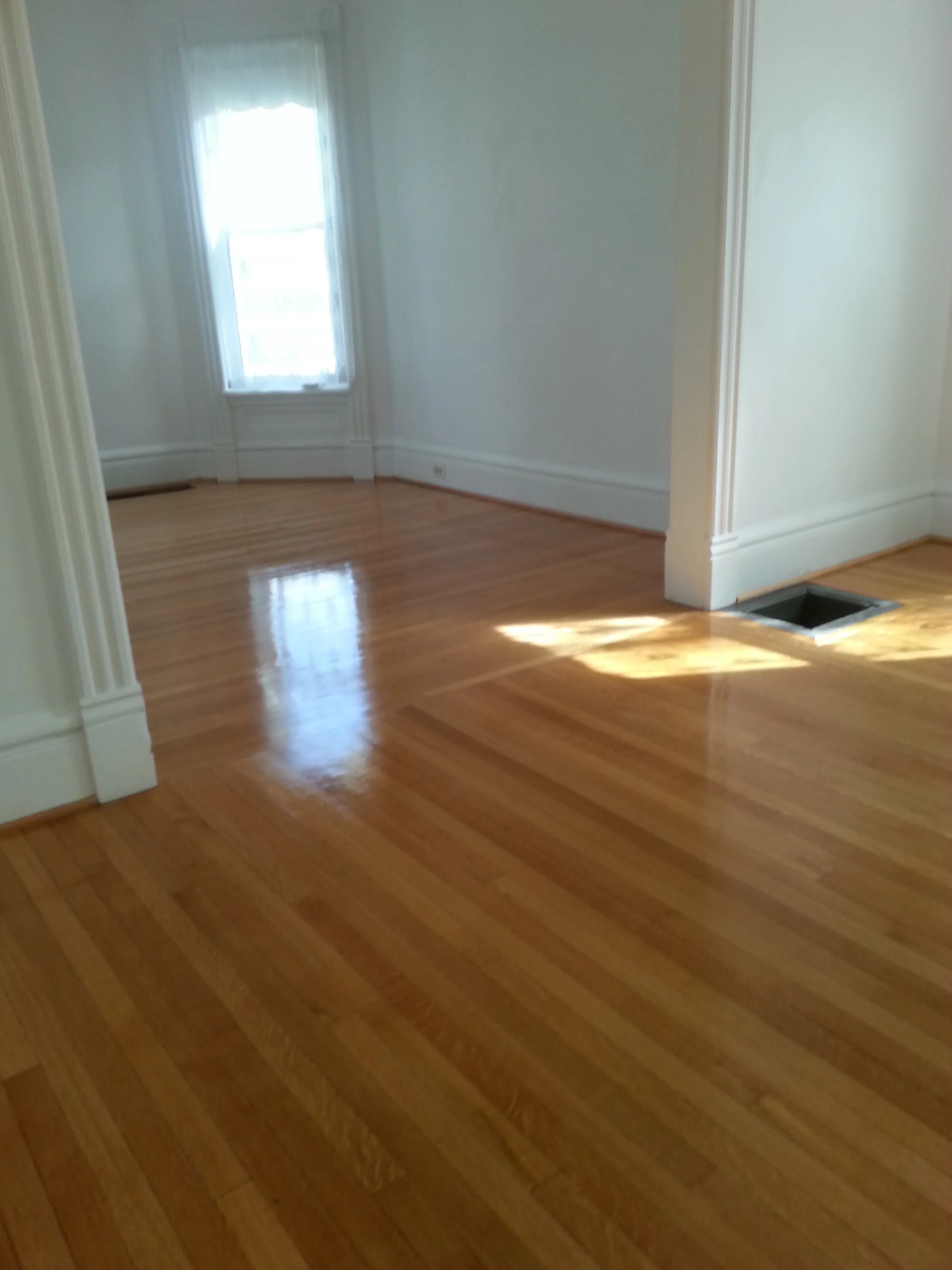Empty room with polished wooden floors, white walls, a large window with sheer curtains, and a ceiling vent.