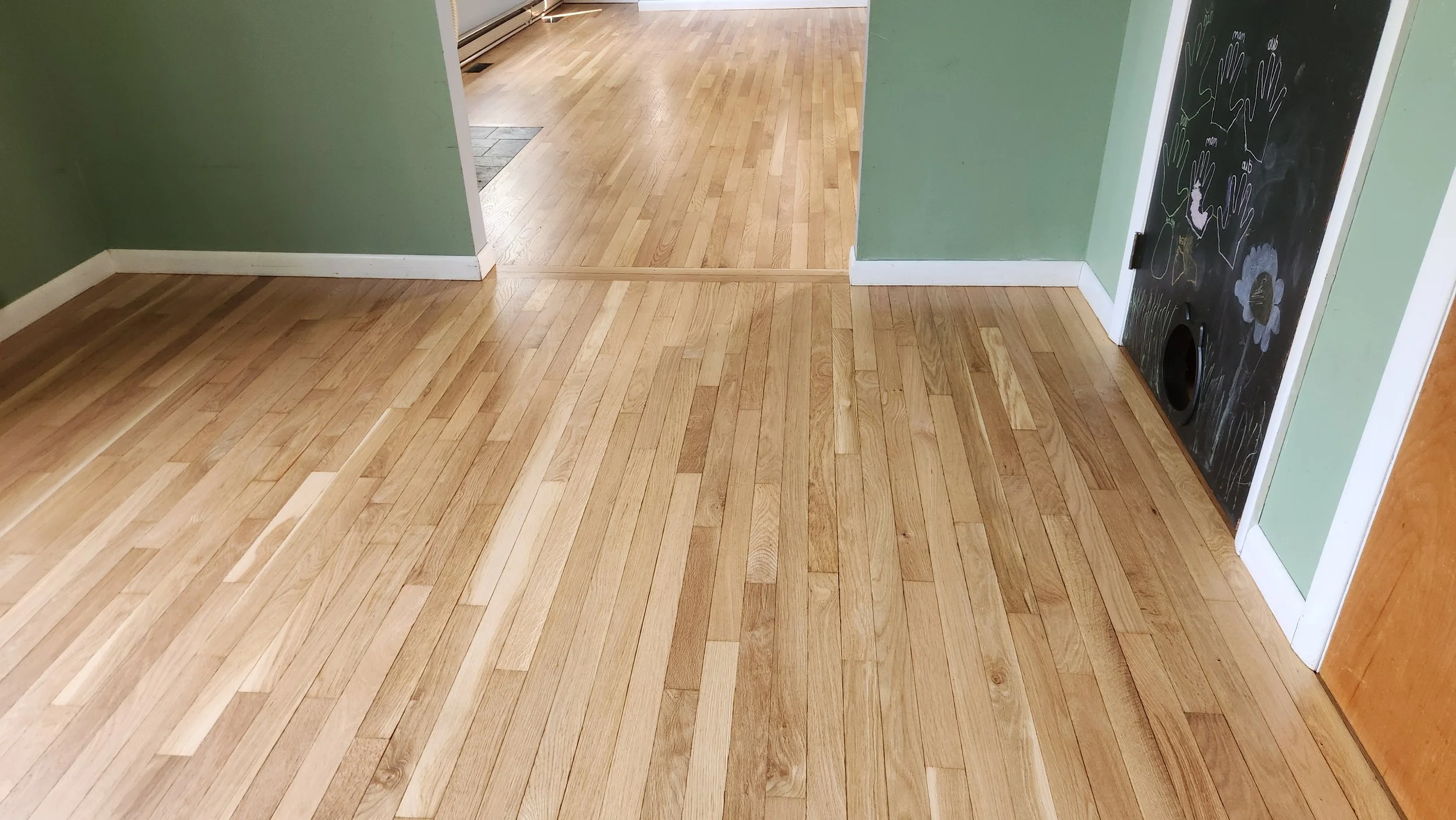 A corner of a room with wooden floors, green walls, white baseboards, and a black chalkboard wall with drawings of flowers and hands.
