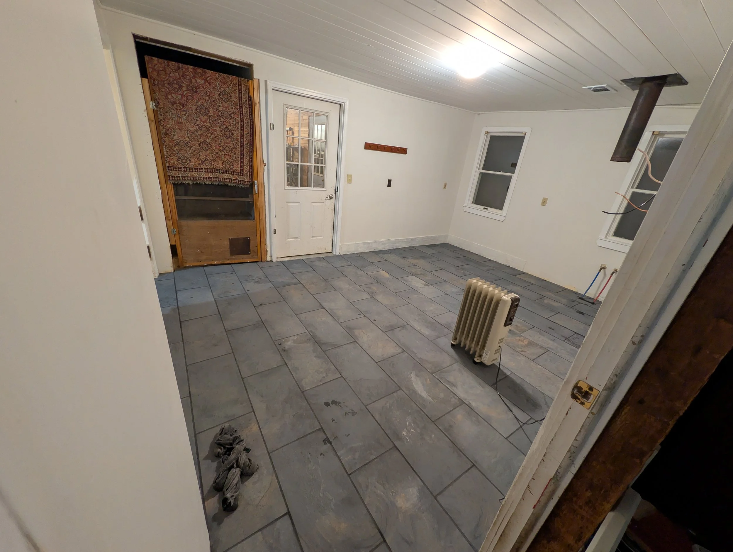 Room under renovation with gray tiled floor, white walls, two windows, an electric heater, and an unfinished interior door with a carpet covering part of the door frame.