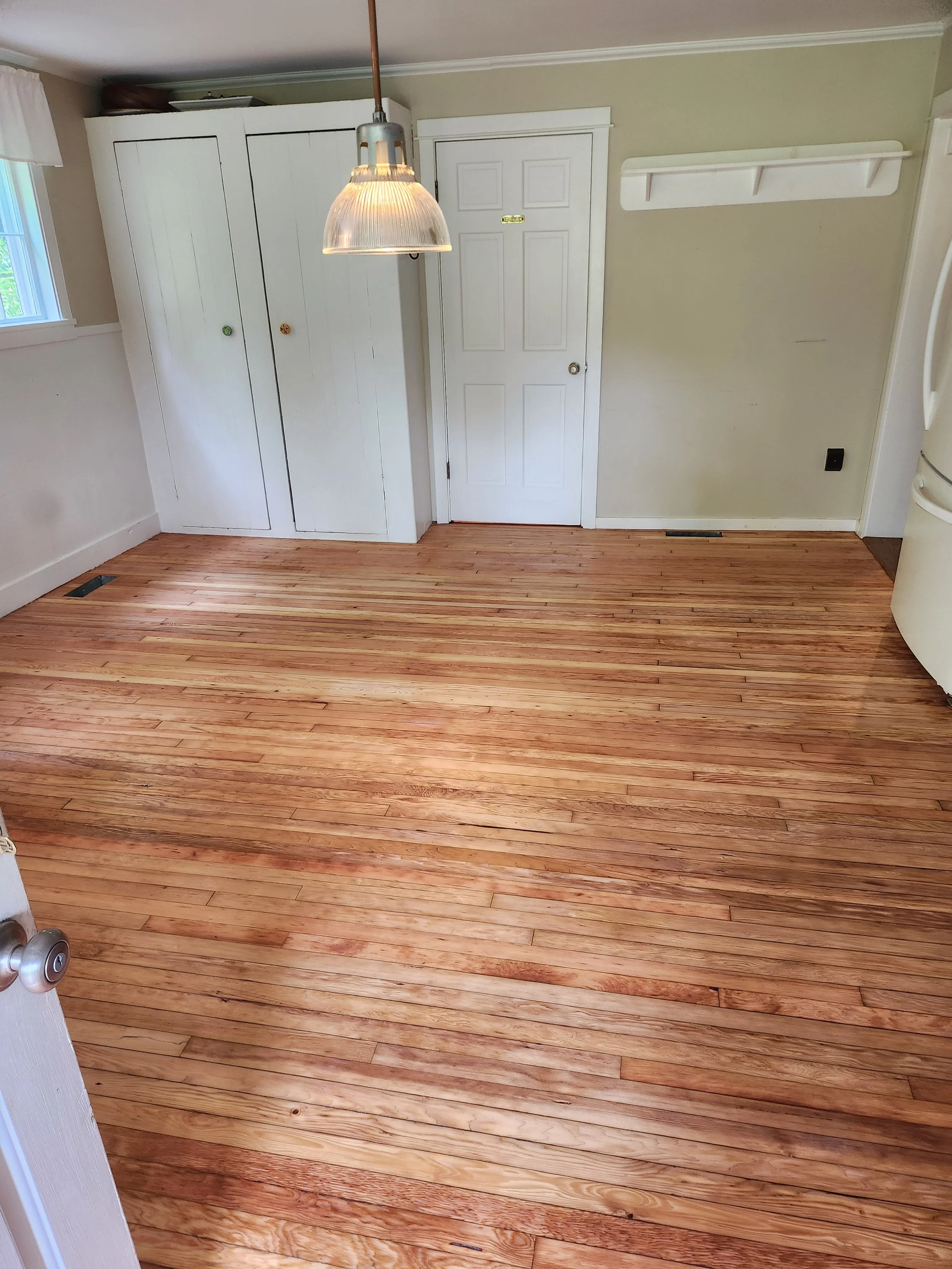 Empty kitchen or dining room with freshly sanded hardwood floors, white cabinets, a white door, and a pendant light hanging from the ceiling.