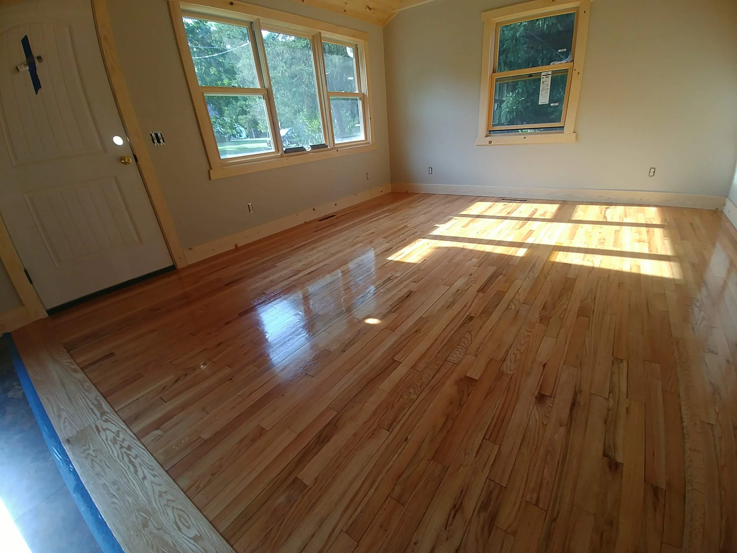 A room with newly finished hardwood flooring and large windows letting in sunlight, showing a partially finished interior space.