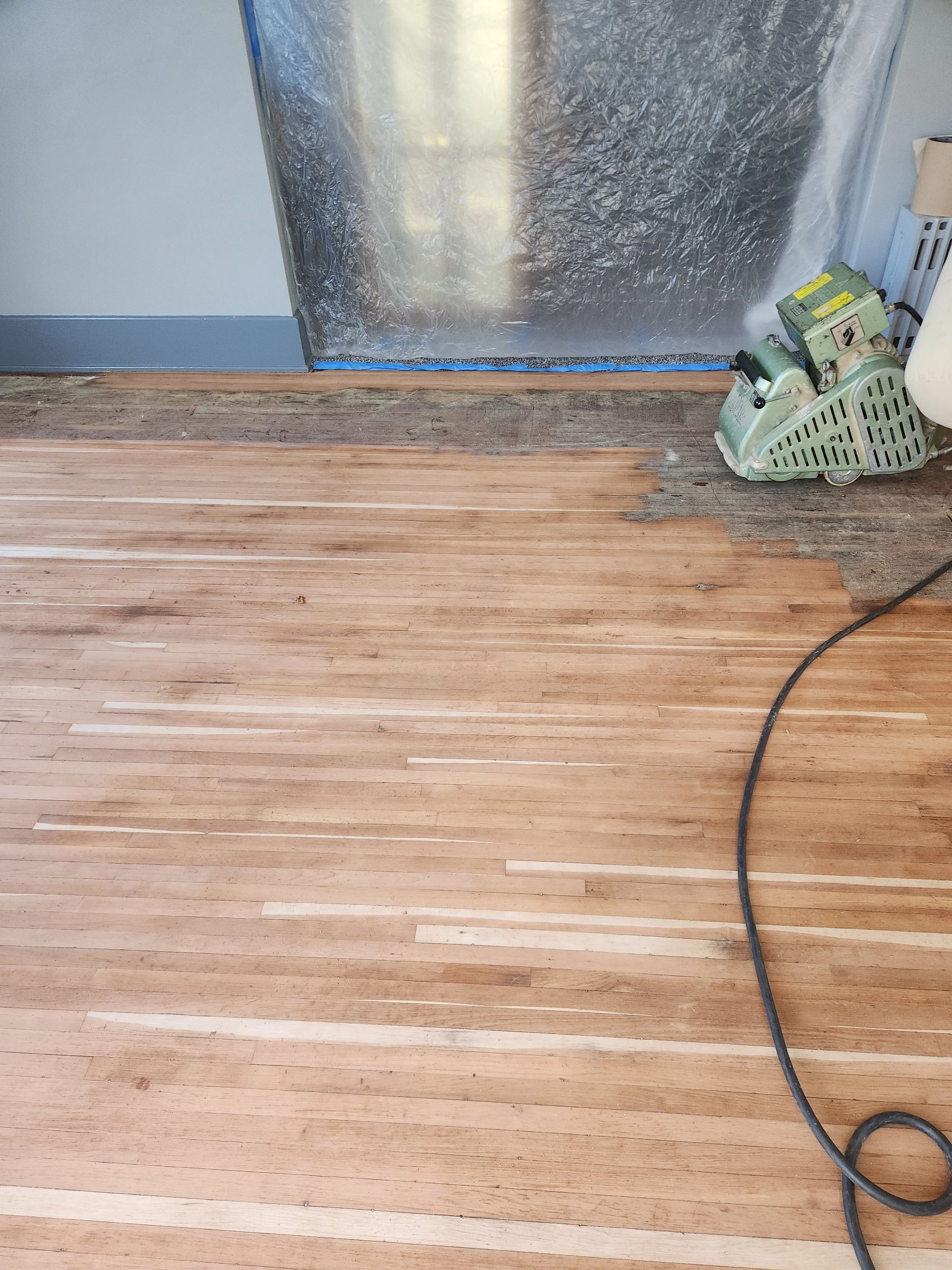 A partially sanded hardwood floor with a floor sander machine nearby and a plastic sheet hanging in the background.
