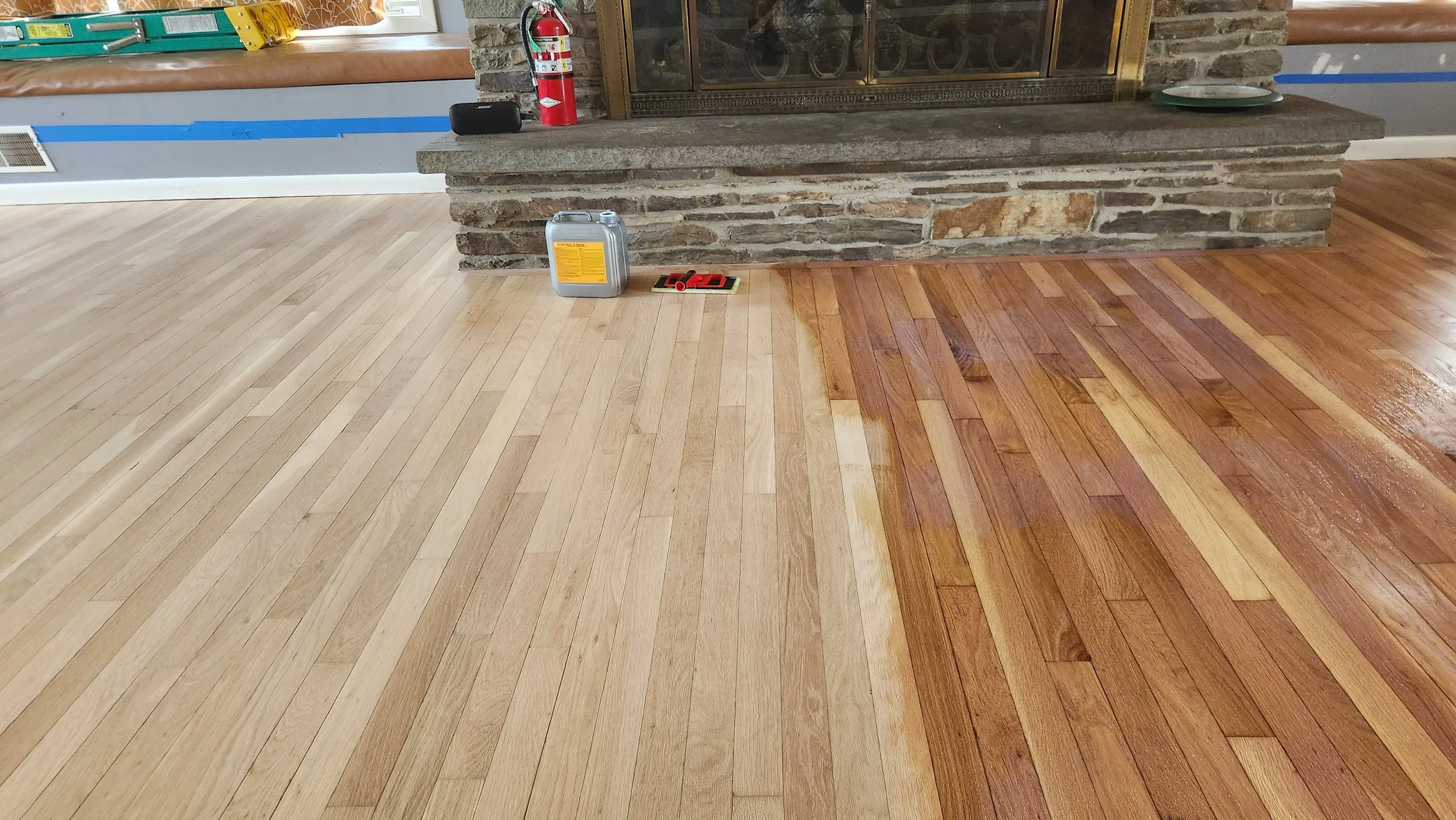A room with a fireplace and a partially finished hardwood floor, with light wood planks on the left side and darker wood planks on the right side. There are tools and items placed on the floor near the fireplace, including a fire extinguisher, contai