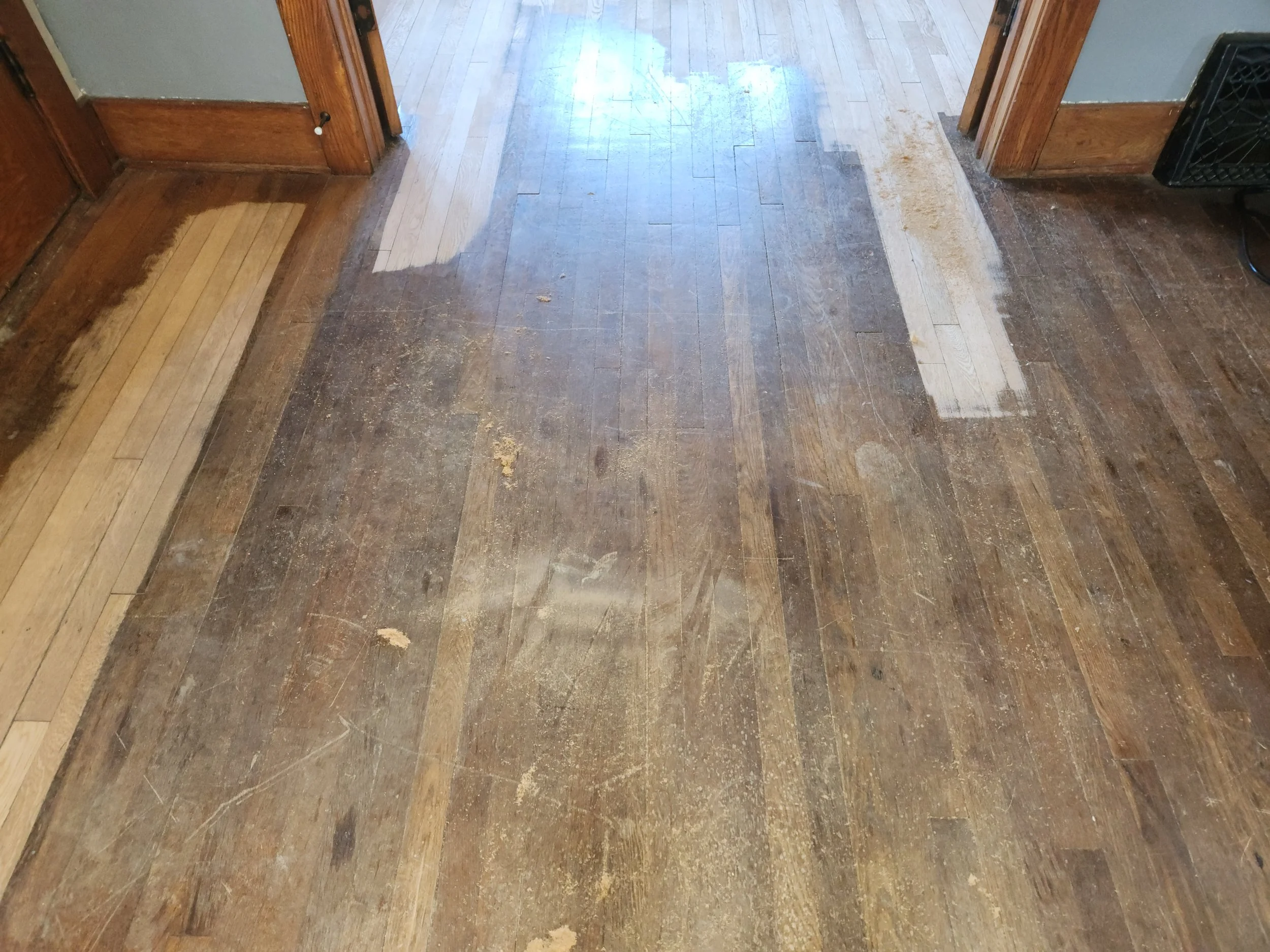Section of a wooden floor with areas of worn, scratched, and partially stripped finish. The middle part is lighter with patches of exposed wood, surrounded by darker, older finish.