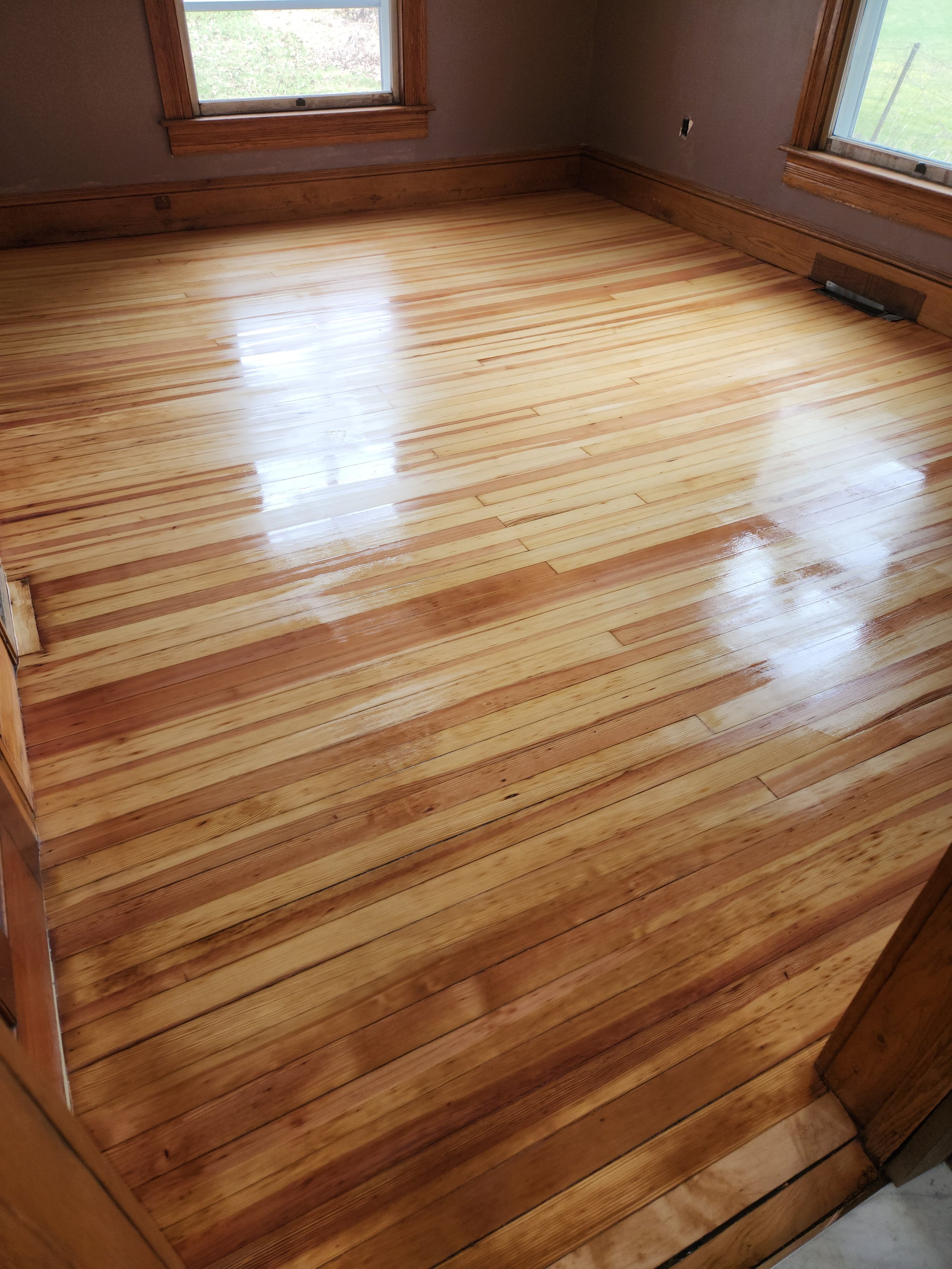 A room with newly polished hardwood floors, two windows allowing natural light, wooden trim around the windows, a vent in the floor, and an empty wall with a hole where a fixture was removed.