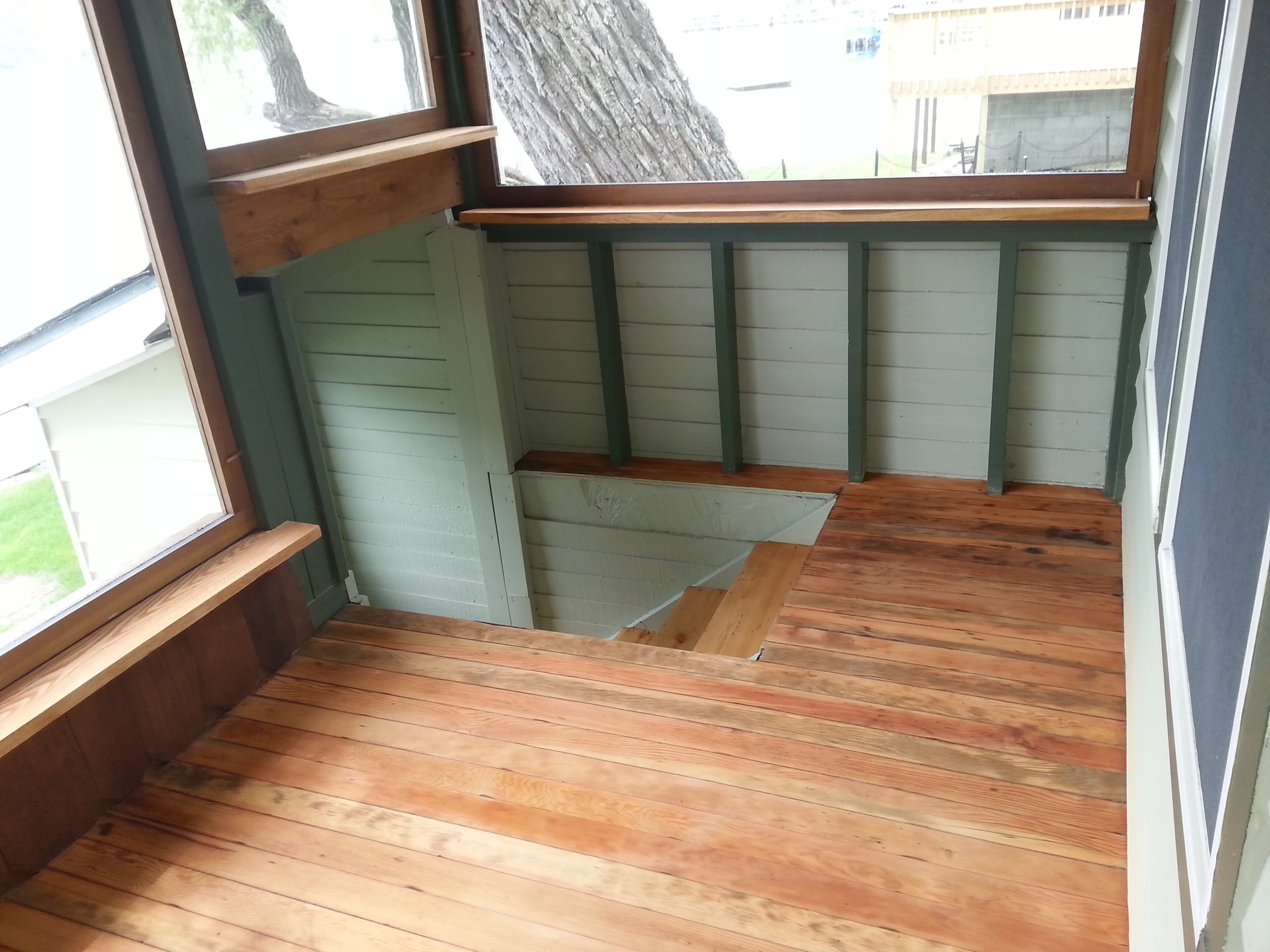 Interior of a small room with polished wooden floors and walls painted in light green. There are large windows on two sides letting in natural light, with a tree visible outside. The room appears to be under construction or renovation, with a stairca
