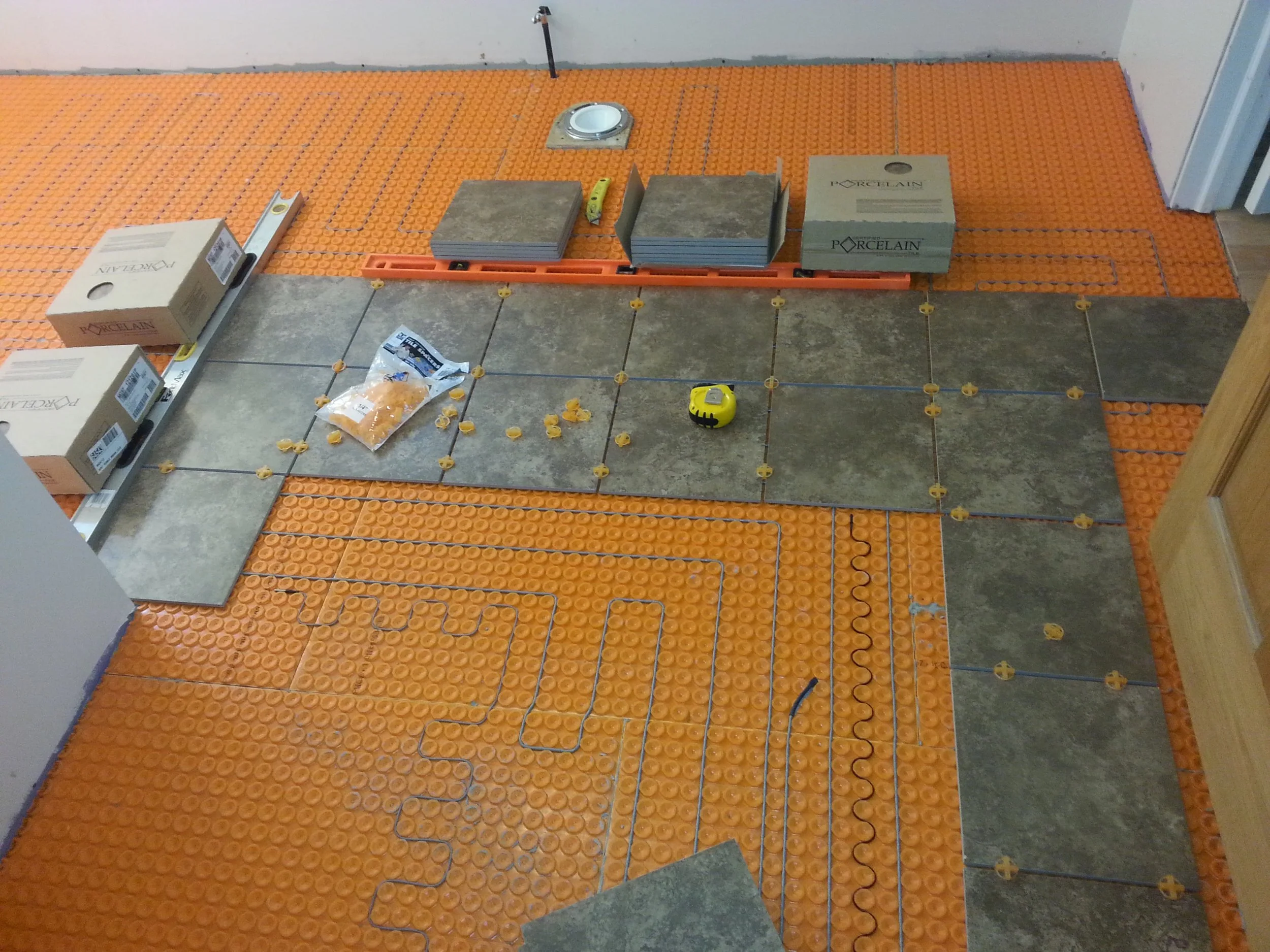 Floor being tiled with gray tiles, orange anti-fracture membrane, and subfloor. Tile spacers and tools are visible, along with boxes of tiles.