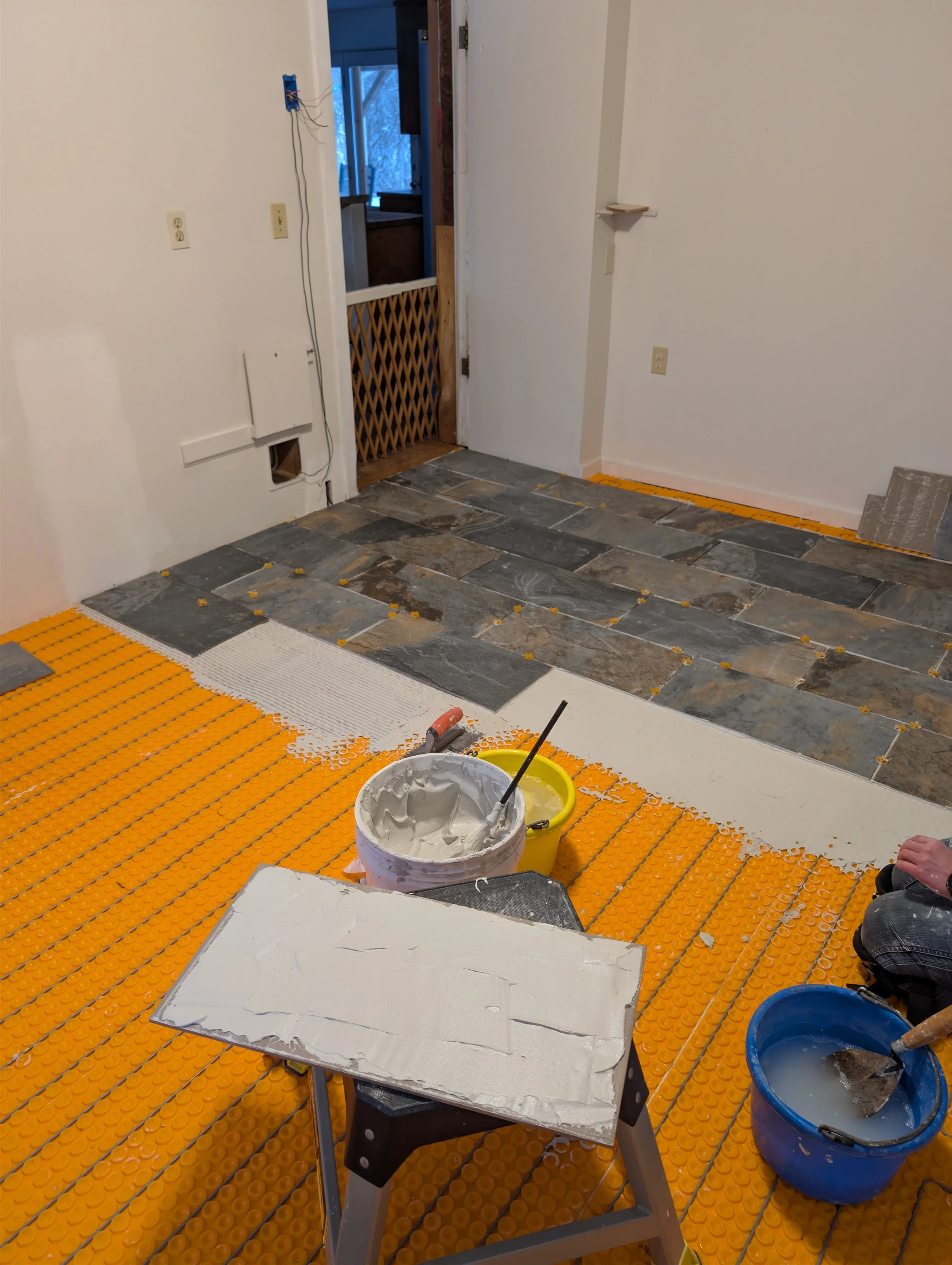 Room under renovation with orange tile floor being installed, buckets of adhesive and tools, doorway leading outside, part of wall with electrical outlets, construction materials and traffic cones present.