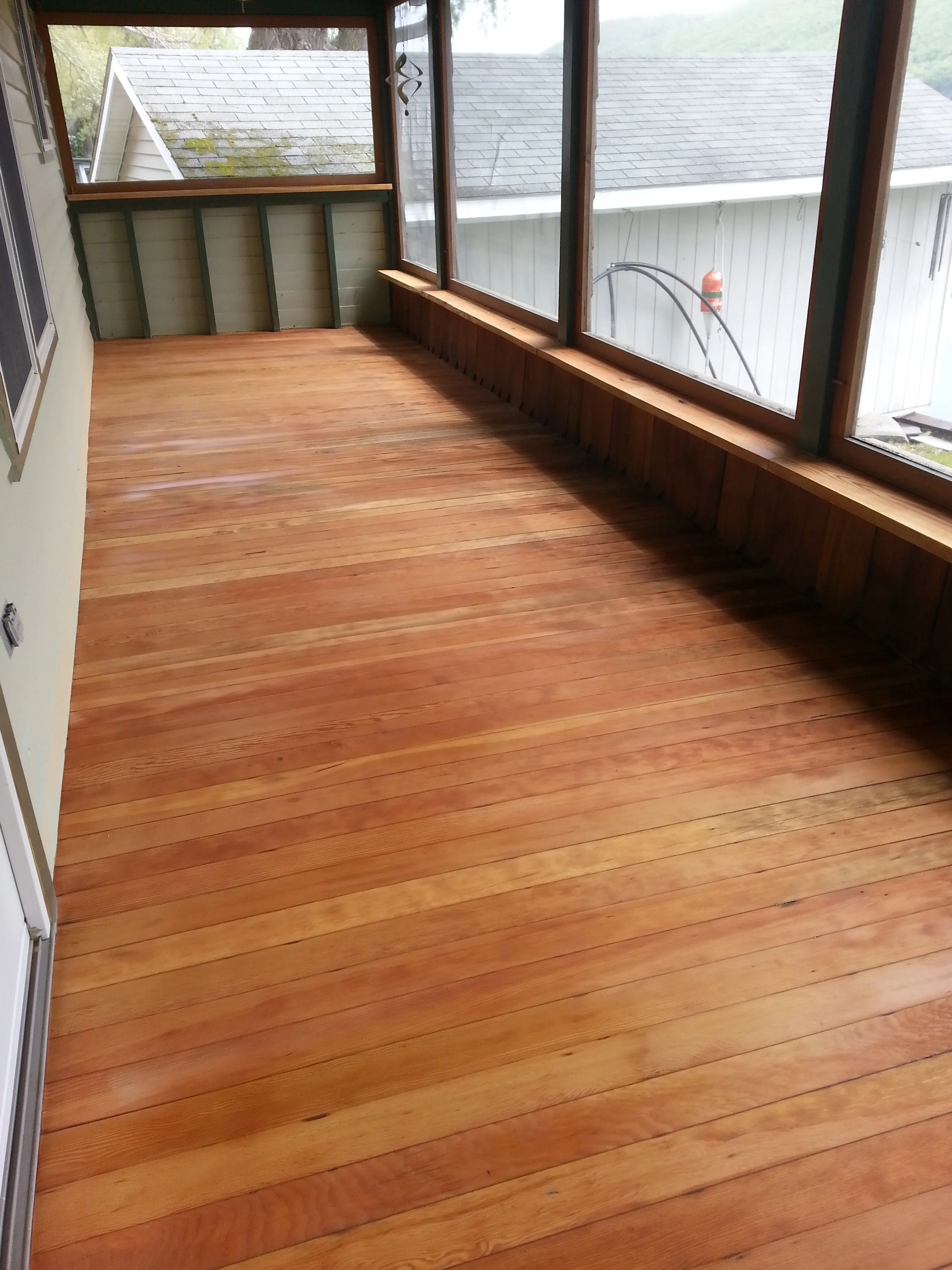 A sunroom with newly refinished wooden floorboards and large windows with a view of neighboring houses and greenery.