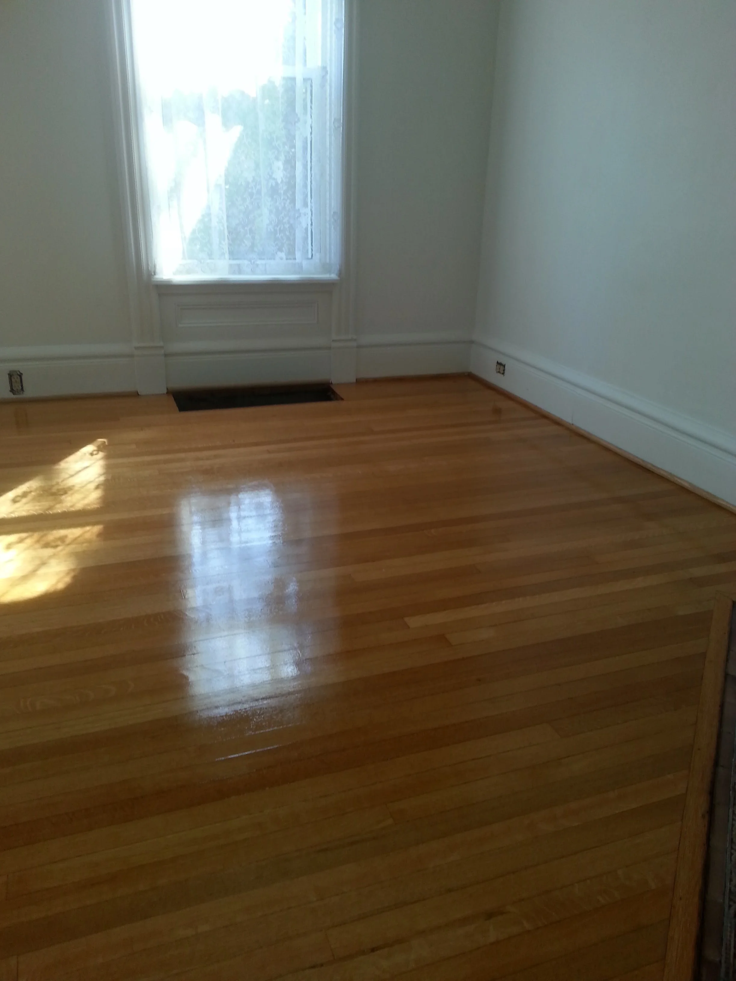 A clean, empty room with hardwood floors, a window with curtains, and a vent near the baseboard.