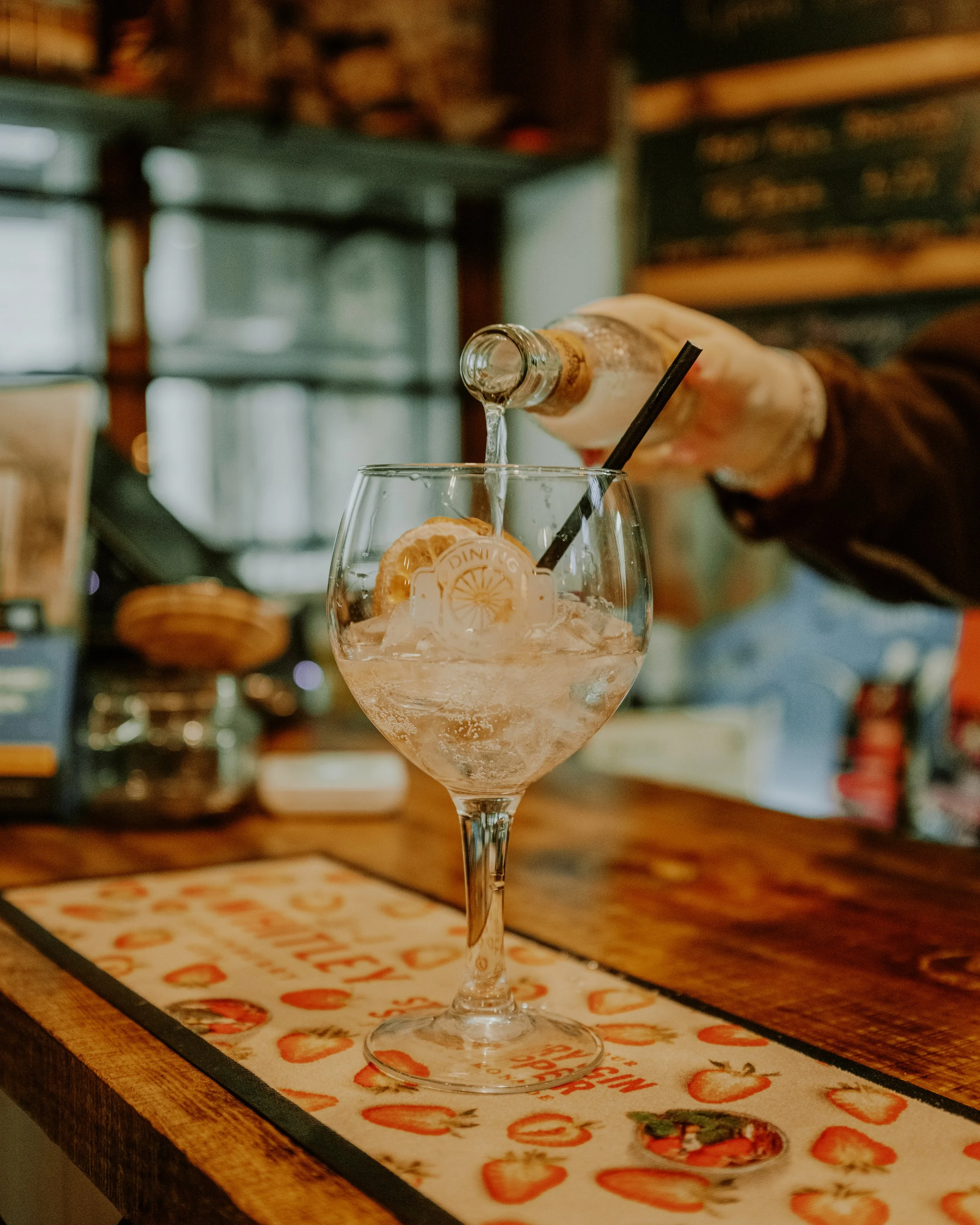 A person is pouring a clear liquid into a large wine glass with ice and lemon slices, on a tray with strawberries, in a cozy, rustic bar or cafe setting.