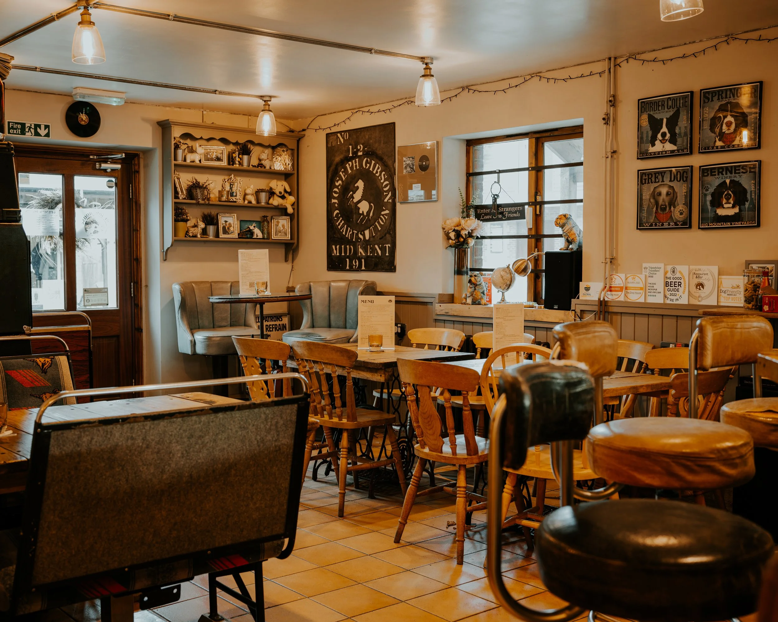 Interior of a cozy pub or restaurant with wooden and cushioned chairs, tables with menus, and wall decorations including framed pictures, posters, and shelves with decorative items. Warm lighting fixtures hang from the ceiling.