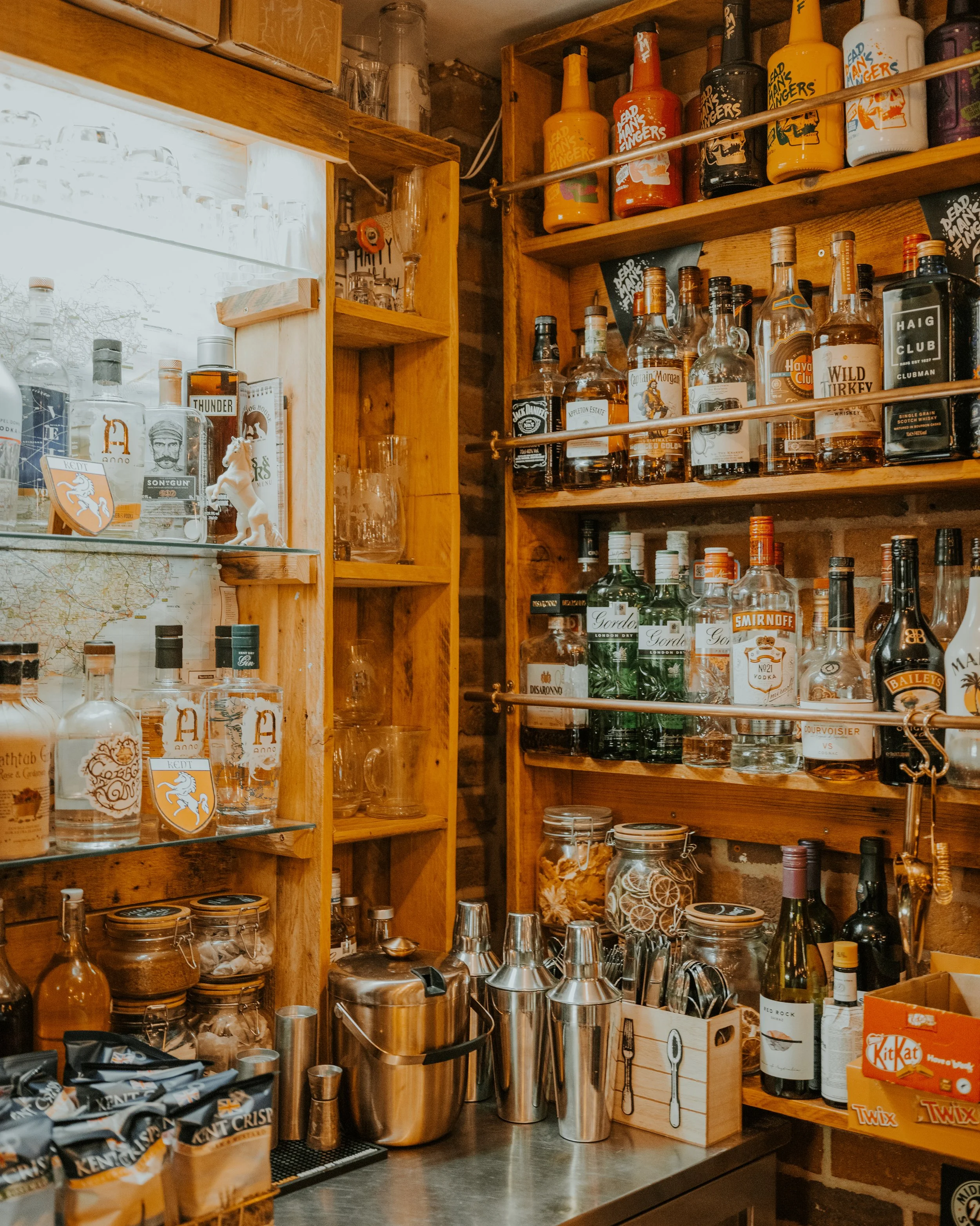 A bar with shelves stocked with various liquor bottles, glasses, and bar tools, including cocktail shakers and jars of ingredients.