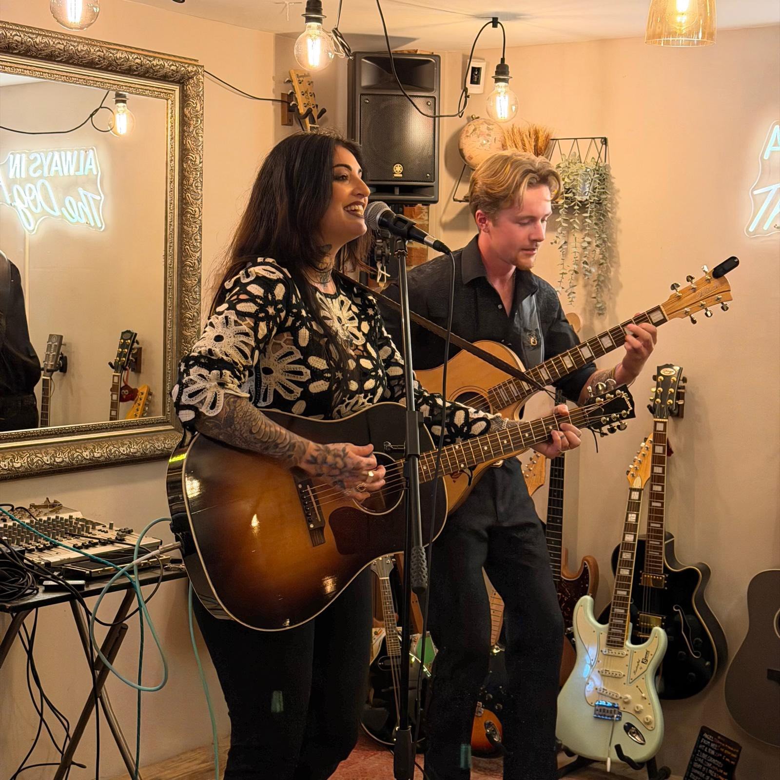 A woman and a man playing guitars and singing at a small indoor music venue, surrounded by various guitars and equipment.