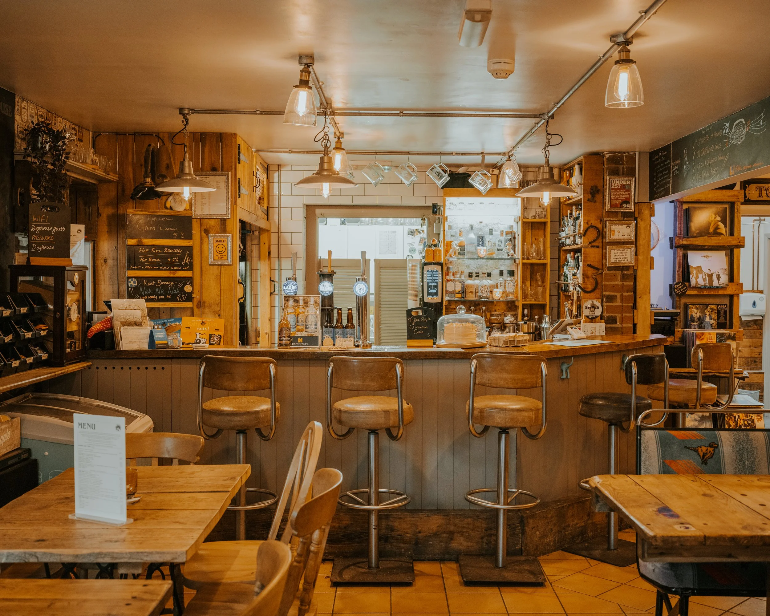 Inside a cozy café with wooden furniture, a bar counter with four barstools, shelves with bottles and snacks, and warm lighting fixtures.