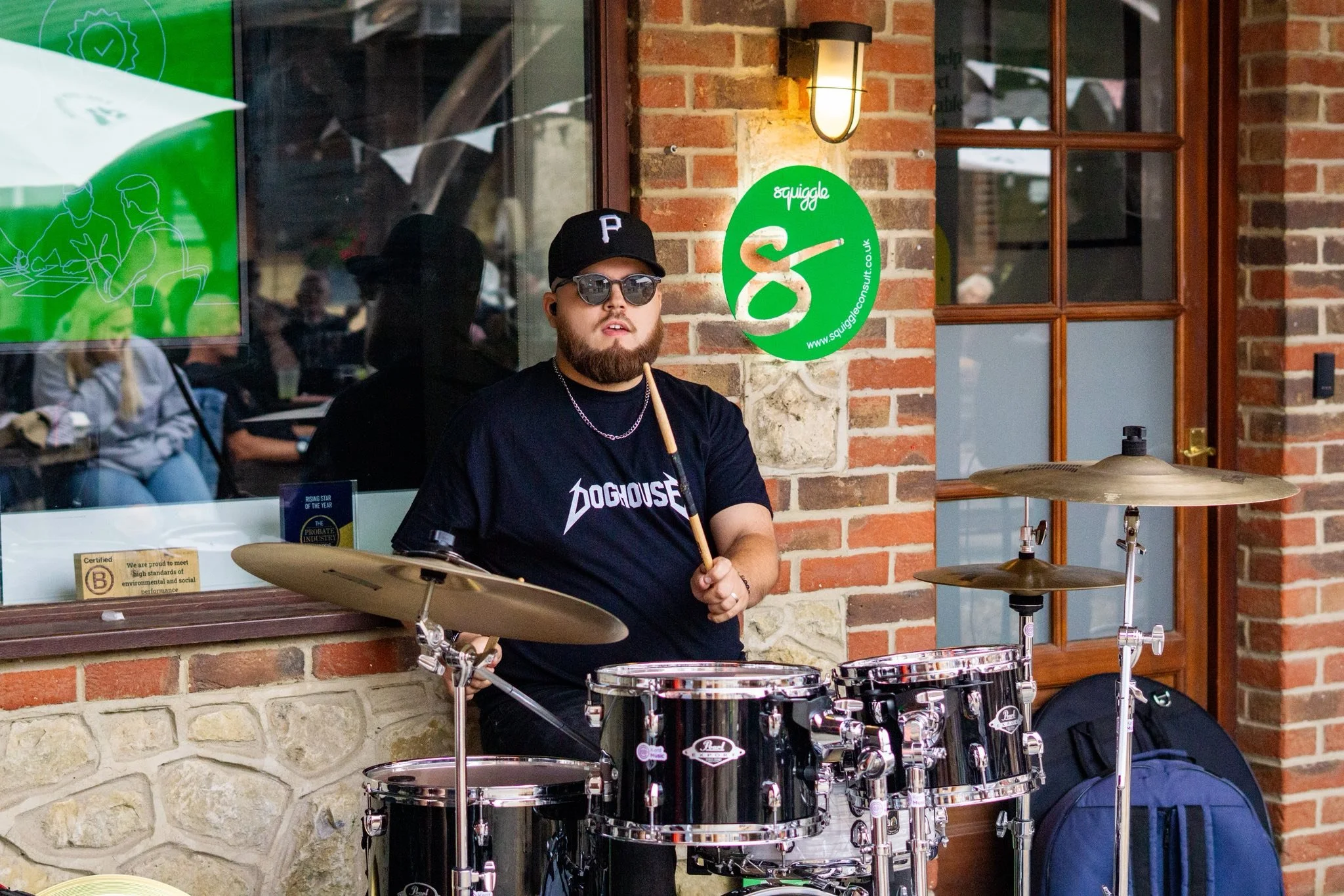 A man with a beard wearing sunglasses, a black cap with a P, a chain necklace, and a black t-shirt with white text, standing next to a drum set outside a building with brick walls. The building window displays a green sign with a snake logo and the w