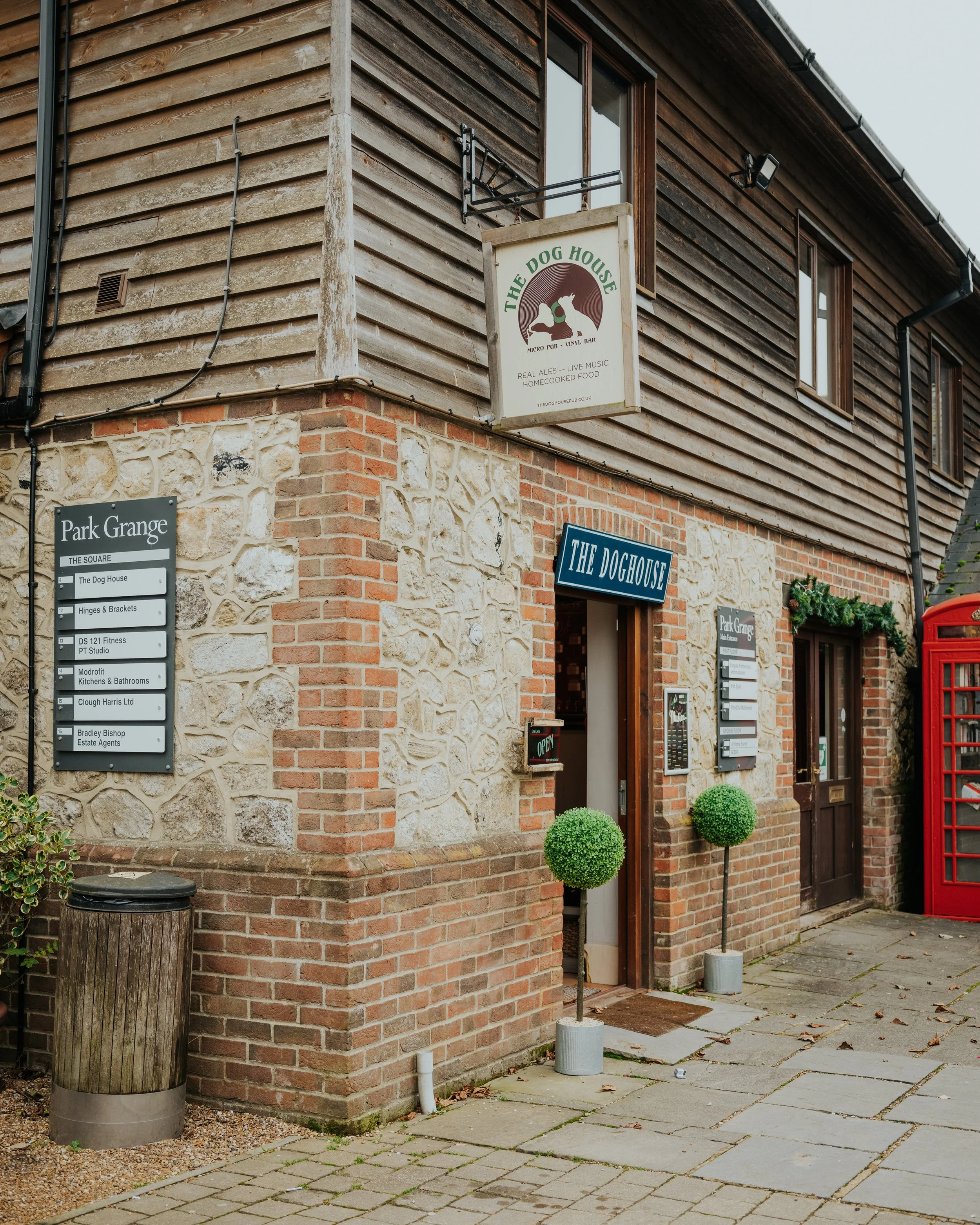 Exterior of a building with signs for The Dog House pub and other businesses, brick and stone facade, two small ornamental trees, a red mailbox, and a sidewalk with leaves.