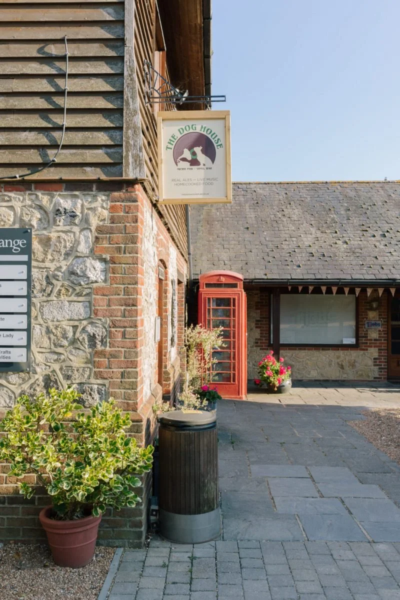 Exterior of a building with a sign reading 'The Dog House,' a vintage red telephone booth, potted plants, and a trash bin.