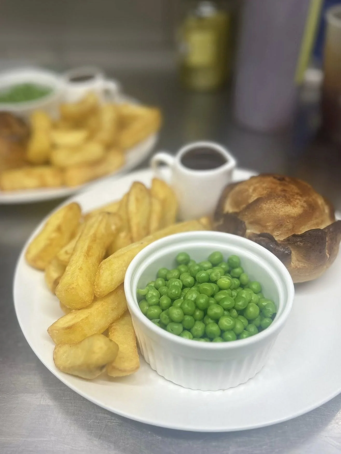 A plate of British breakfast food with chips, green peas, a large baked Yorkshire pudding, and two small containers of brown gravy and dark sauce.