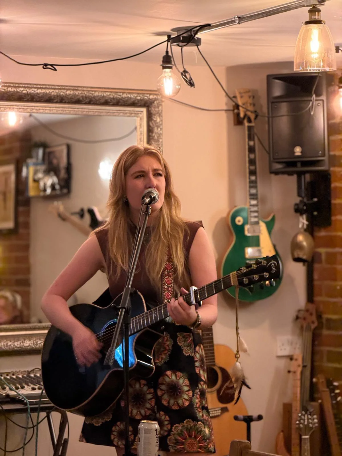 A woman with long blonde hair singing into a microphone and playing an acoustic guitar on stage in a cozy, decorated room with guitars on the wall, warm lighting, and a large mirror.