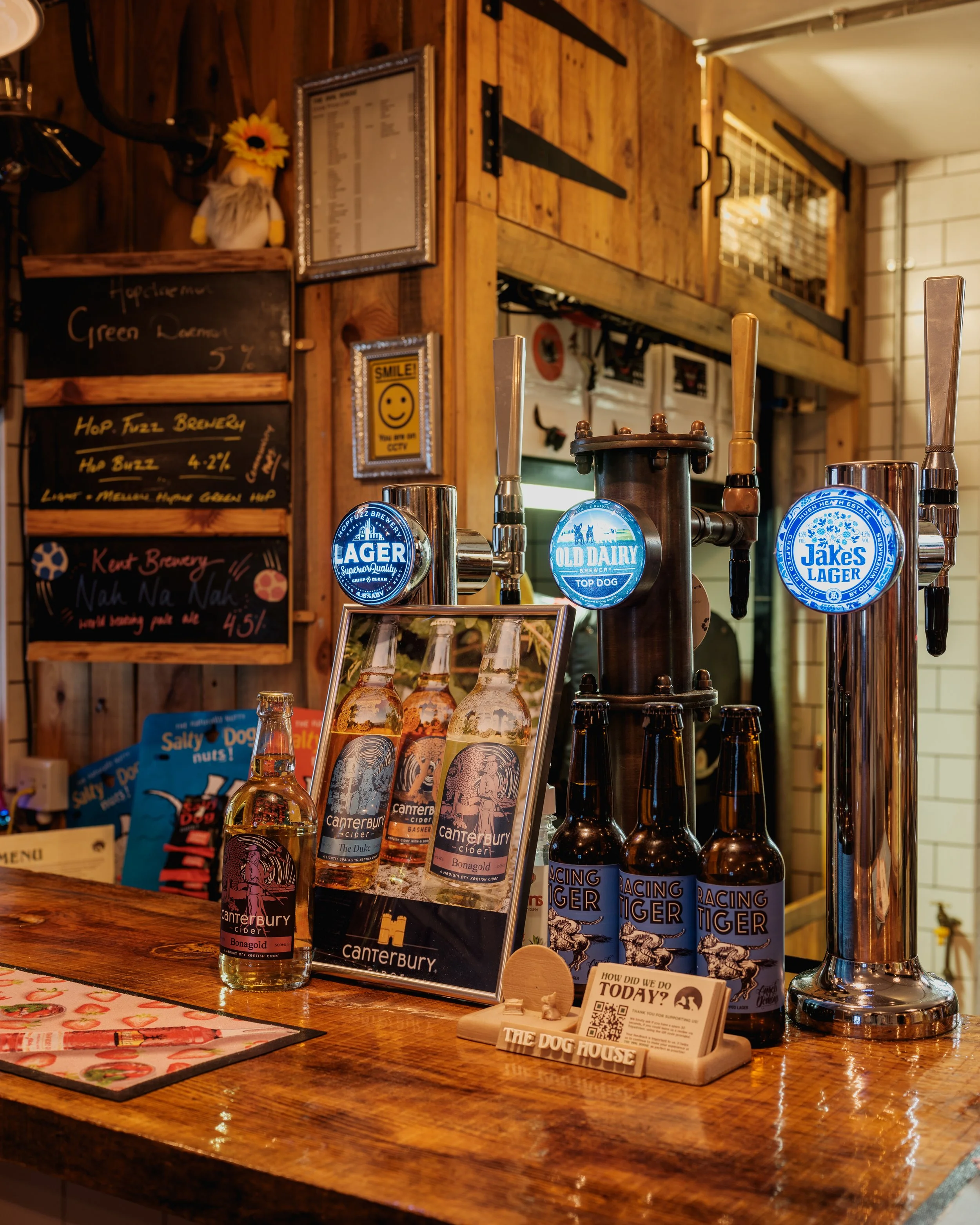 Bar counter with cider bottles, beer taps labeled Lager, Old Dairy, Jake's Lager, and Racing Tiger, and a framed menu, with wood paneling and wall decorations in the background.