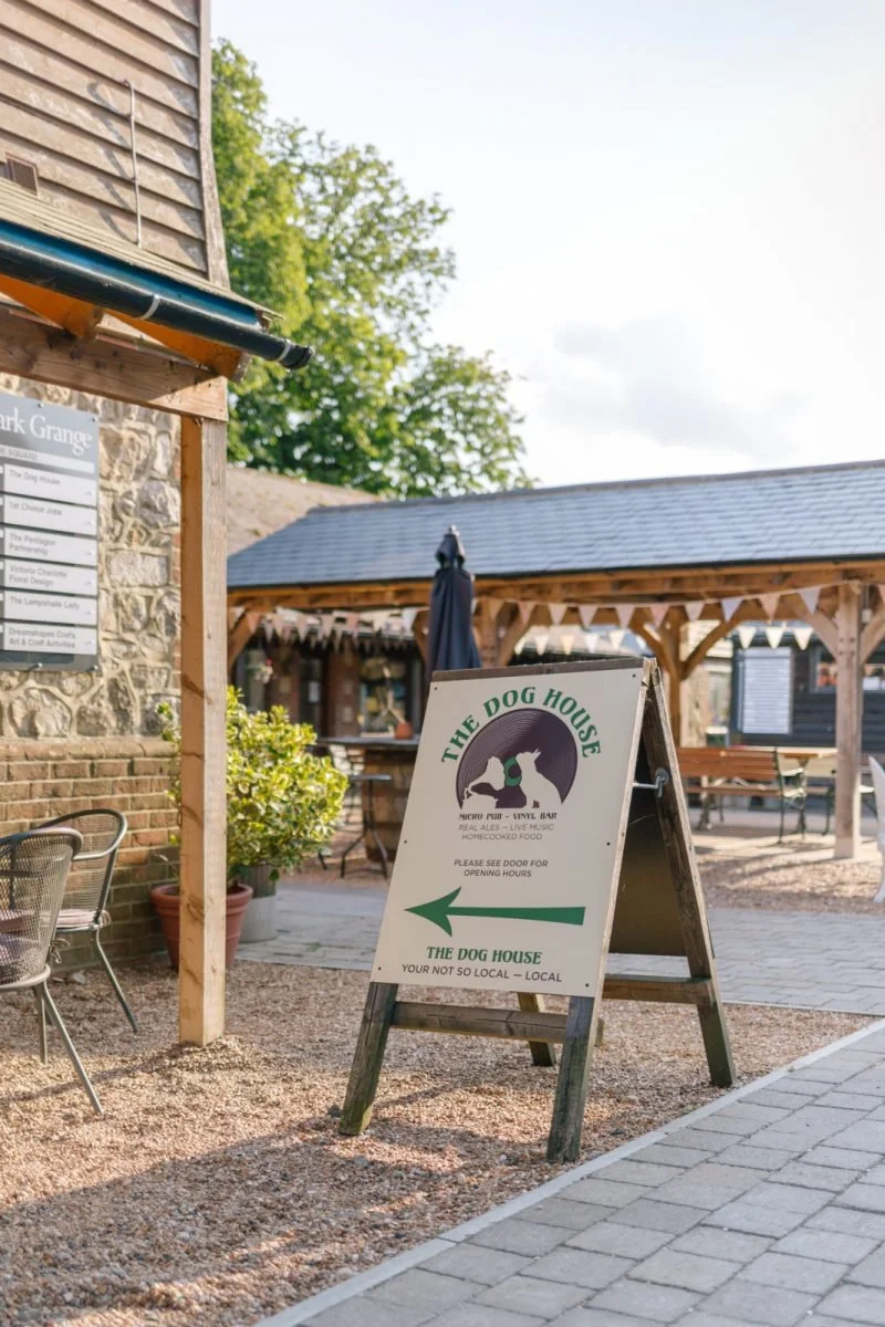 Outdoor signboard for The Dog House, a micro pub and live music venue, with an arrow pointing left indicating the direction to the entrance, set outside under a wooden pavilion with greenery nearby.
