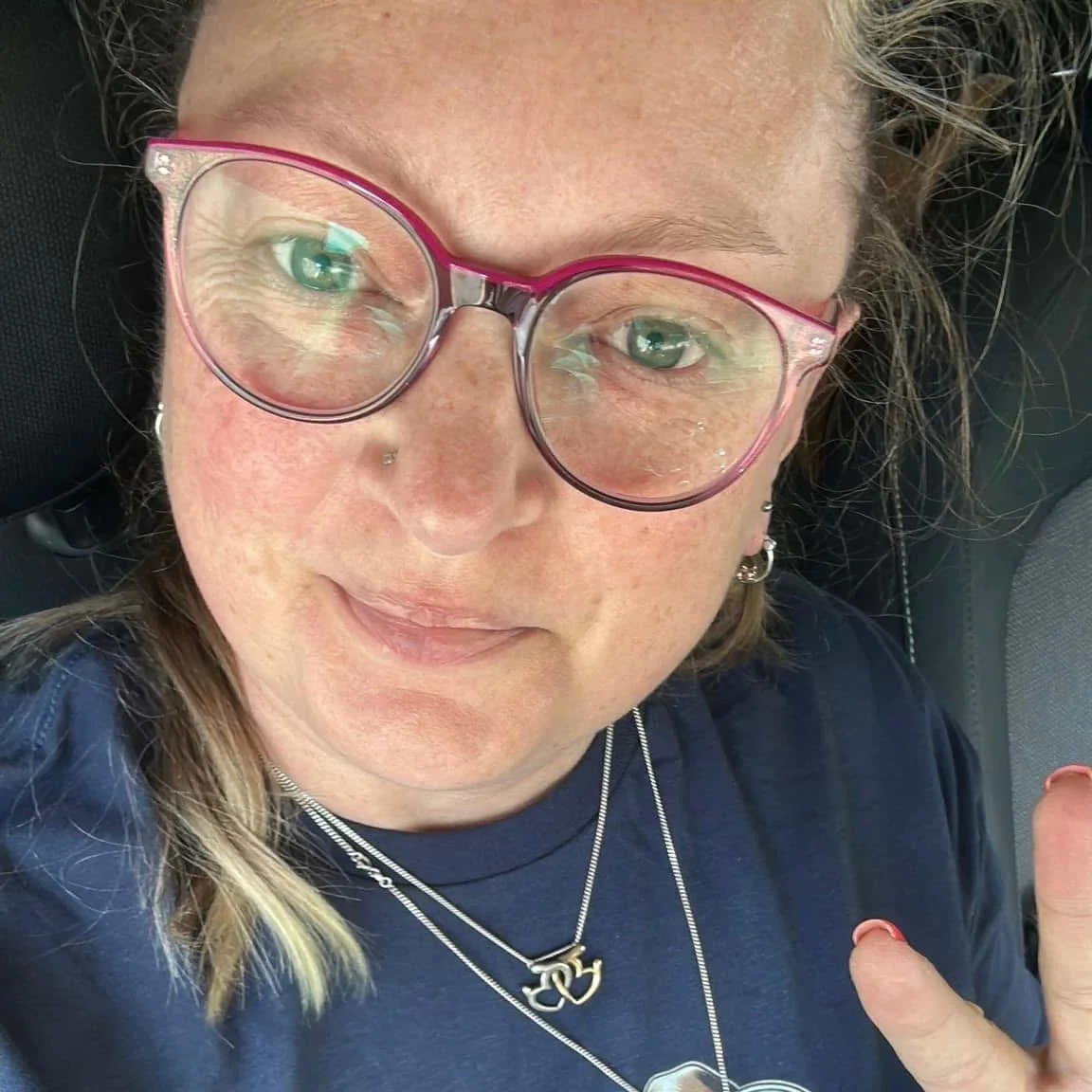 Close-up of a woman with glasses, piercings, and layered necklaces, lying down and smiling.