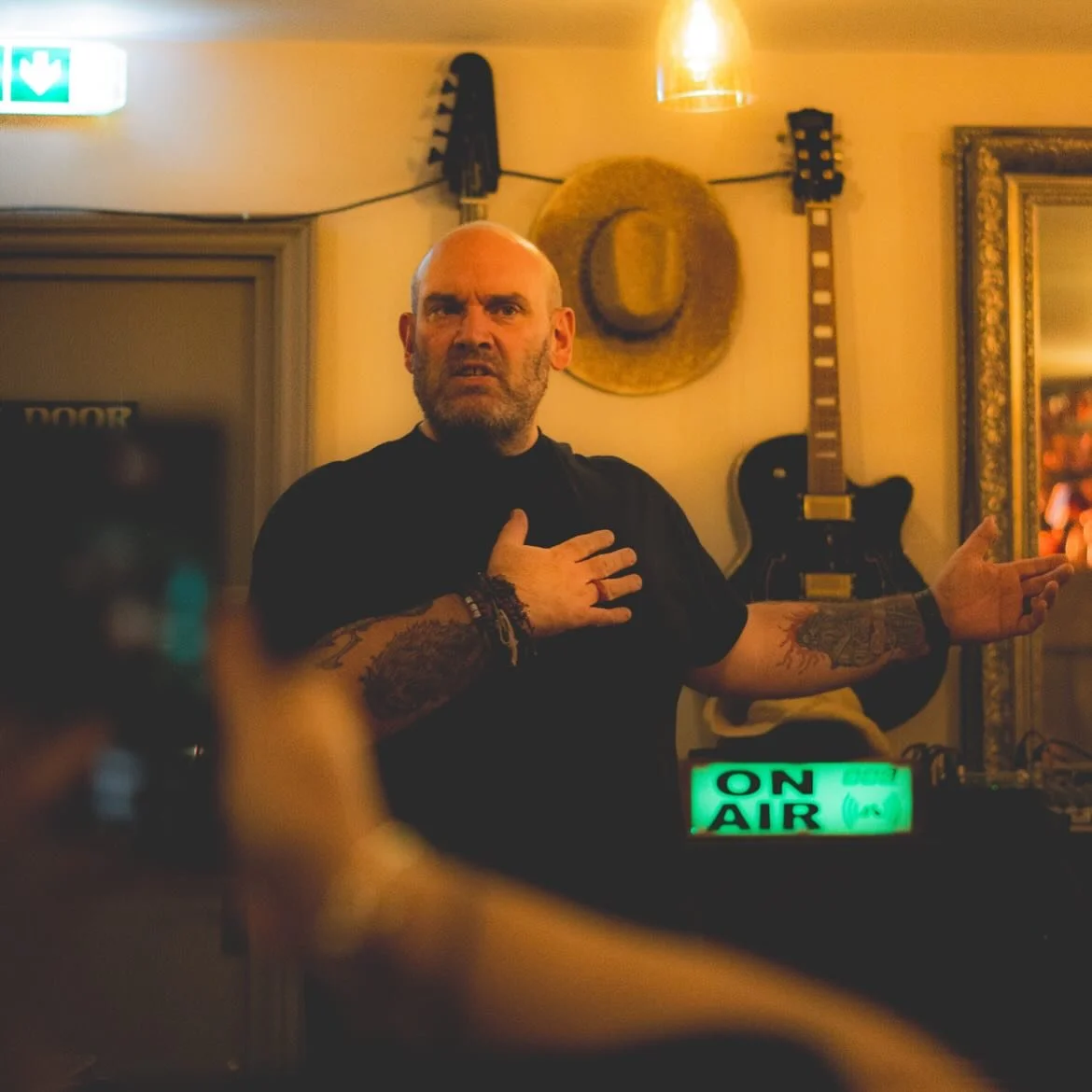 A man with a bald head and tattoos on his arms is gesturing with his right hand on his chest and his left hand extended outward in a dimly lit room decorated with a hat on the wall, a guitar, and a lit