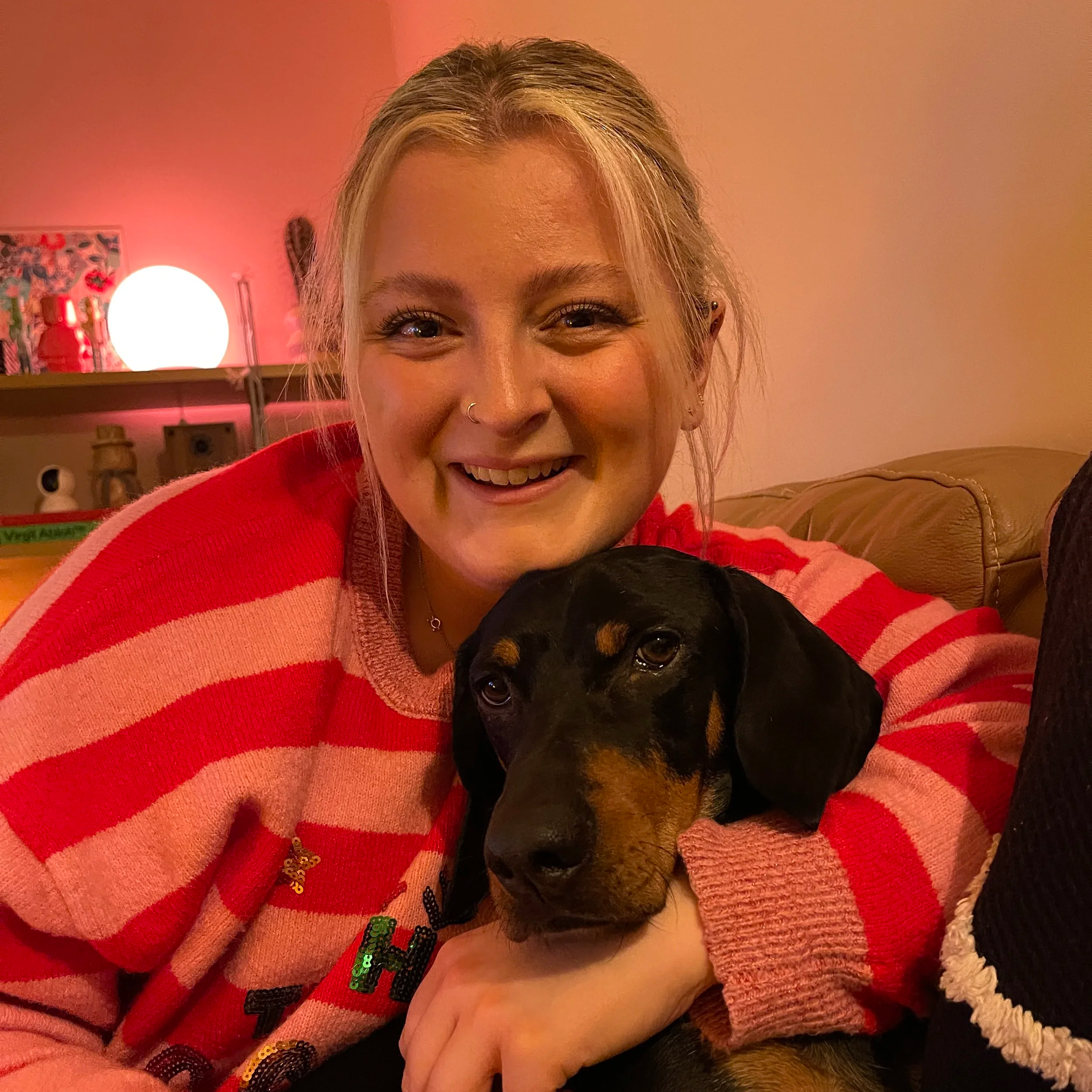 A woman smiling while holding a black and tan Dachshund dog indoors. She is wearing a pink and red striped sweater and the background shows a shelf with a lamp and various decorative items.