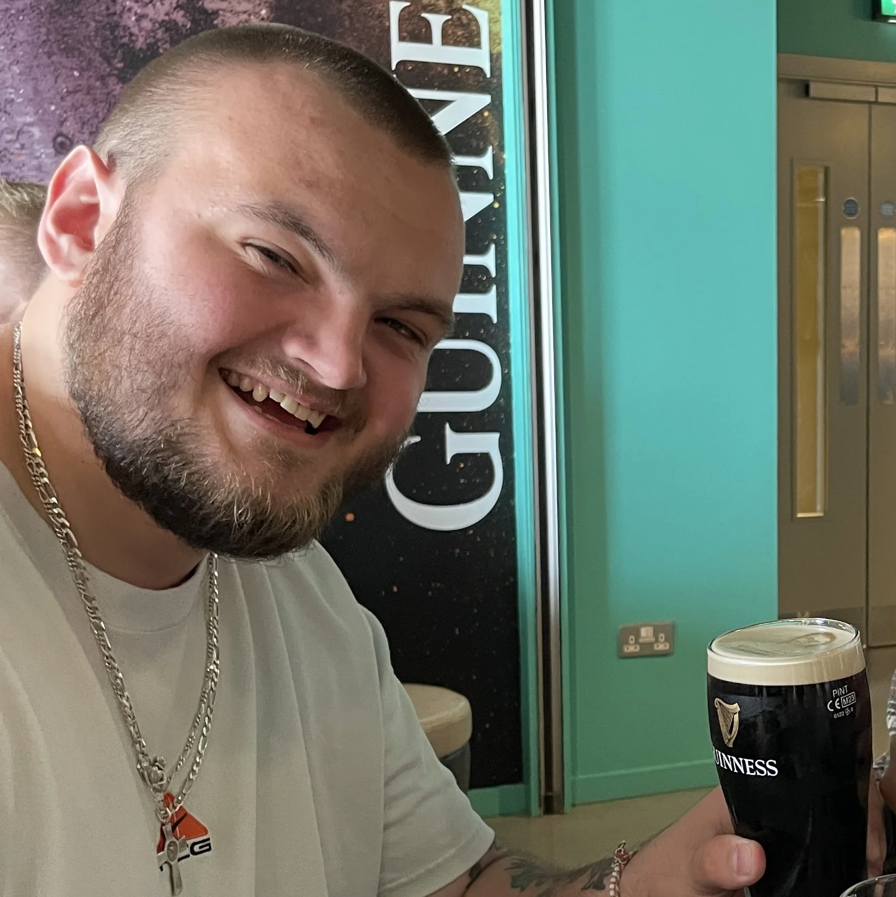 A man with a beard and short hair smiling while holding a pint of Guinness beer in a pub or bar.