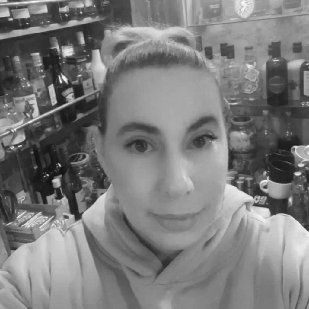 Black and white close-up selfie of a woman with a ponytail, standing in front of shelves filled with bottles and jars.