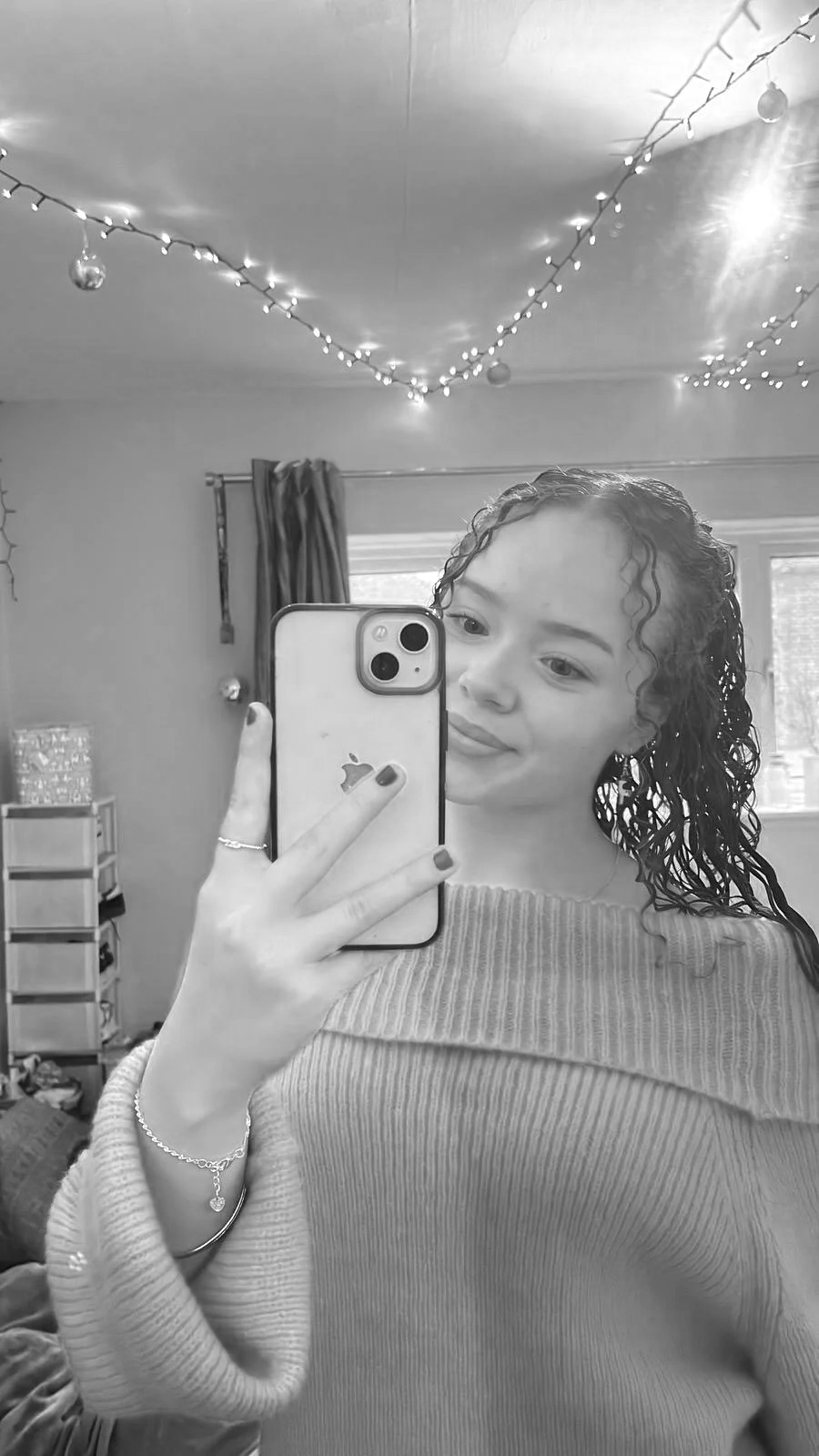 A young woman with curly hair taking a mirror selfie in a room decorated with string lights and ornaments