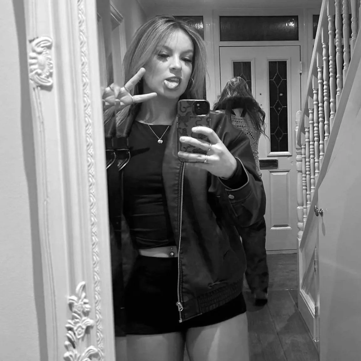 Girl taking a mirror selfie making a peace sign, in a hallway with a woman in background, all in black-and-white.