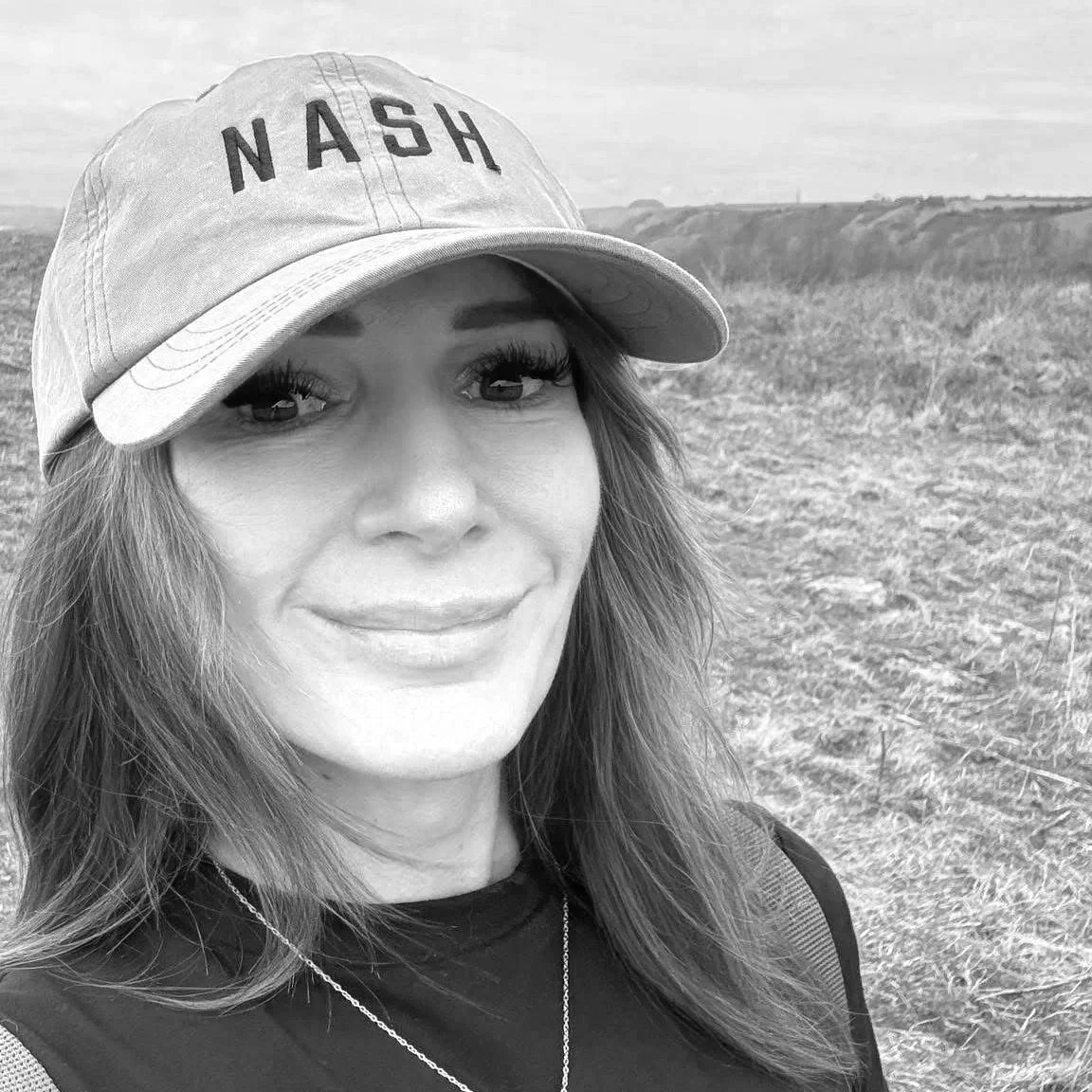 A woman wearing a baseball cap with the word 'NASH' on it, standing outdoors in a grassy field with a distant horizon and cloudy sky in black and white.