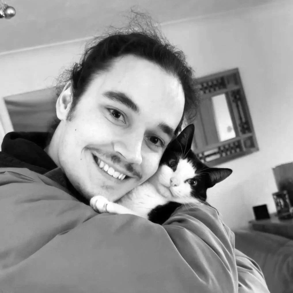 A young man with dark hair smiling and hugging a black and white cat in a cozy living room.