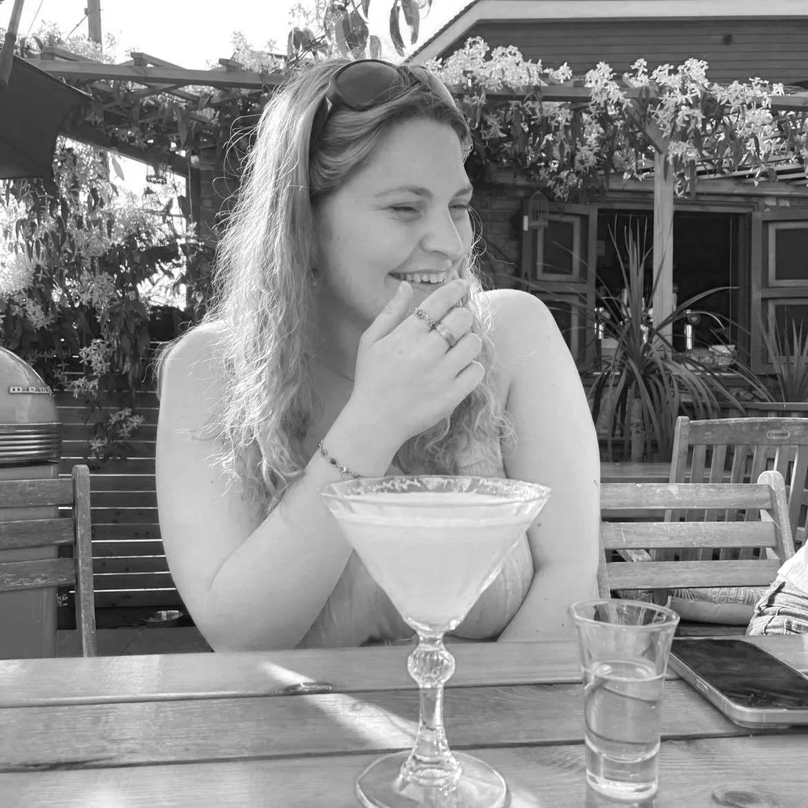A woman smiling and sitting at a table outdoors with a cocktail glass in front of her, wearing sunglasses on her head, in a garden or patio setting.