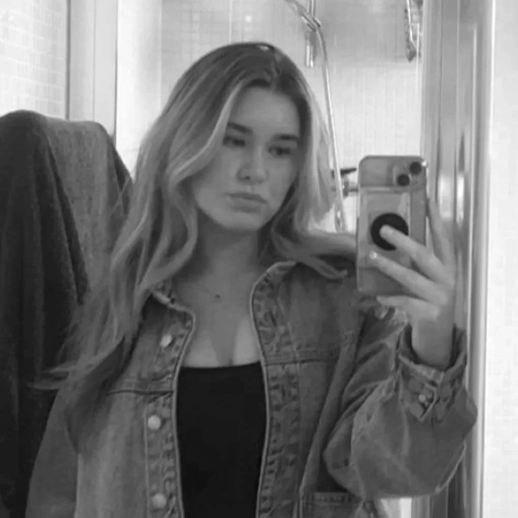 A young woman with wavy hair takes a mirror selfie in what appears to be a bathroom. She is wearing a denim jacket over a black top.