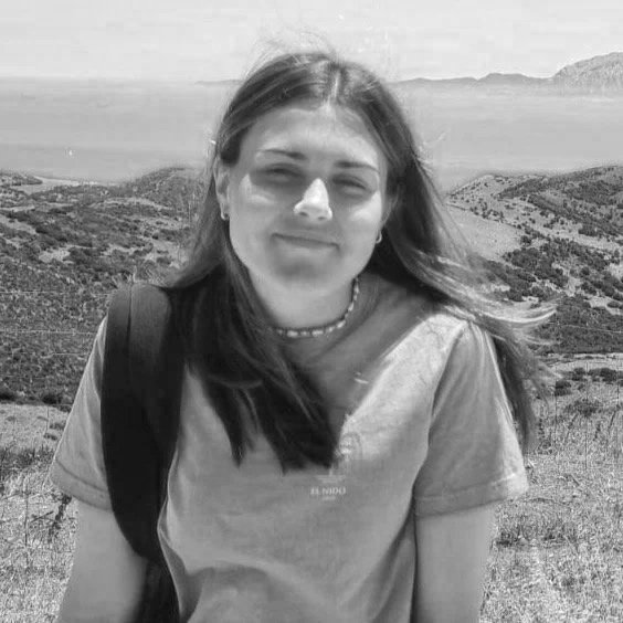 A young woman with long hair standing outdoors in a hilly landscape with water and mountains in the background.