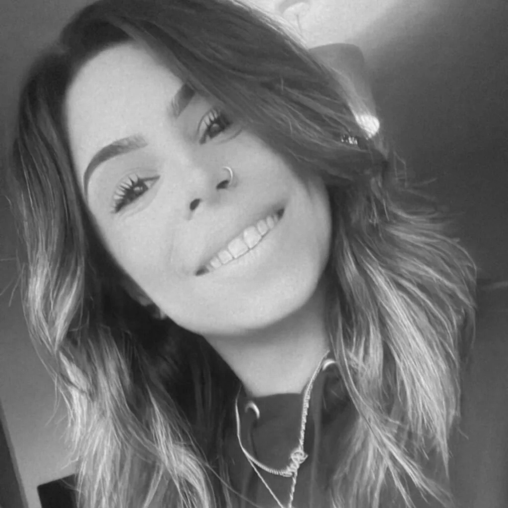 Close-up black and white portrait of a smiling woman with wavy hair, wearing a nose ring and layered necklaces.