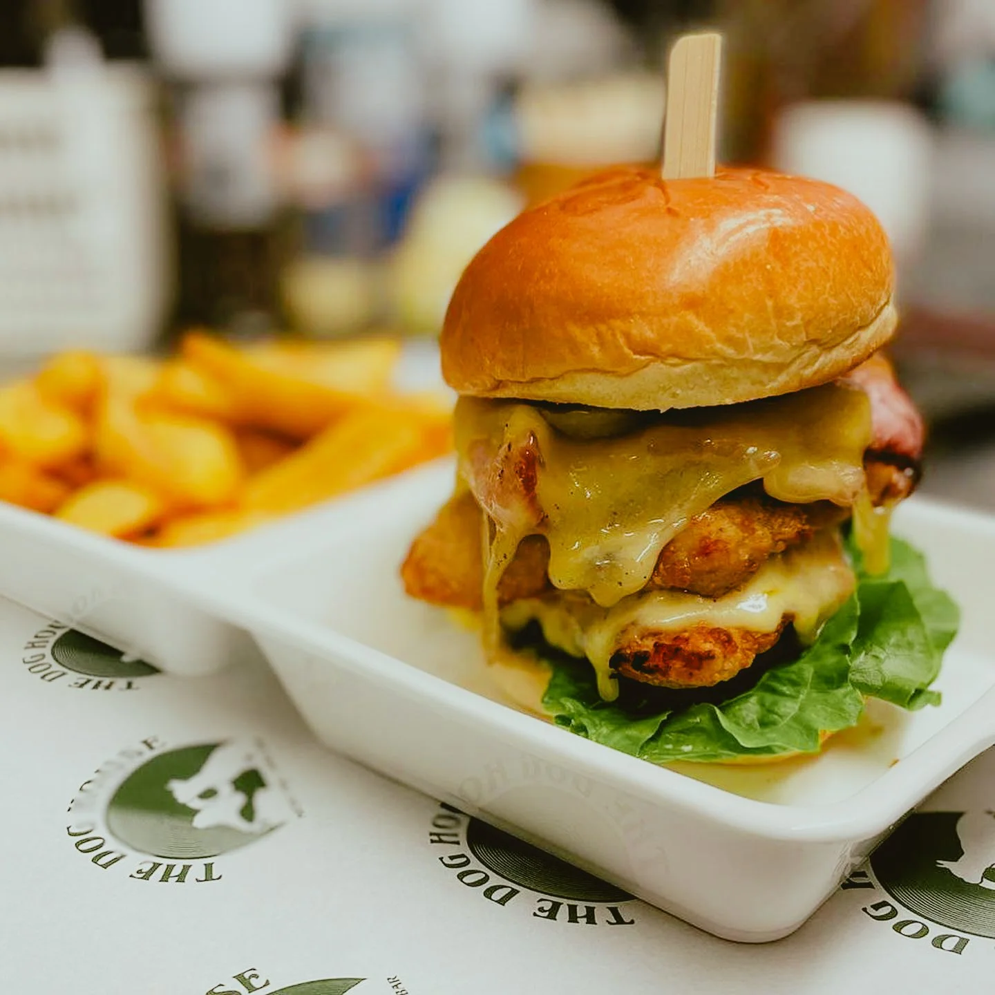 Cheeseburger with lettuce, melted cheese, a fried chicken patty, pickles, onions, and a bun served with French fries on a tray.