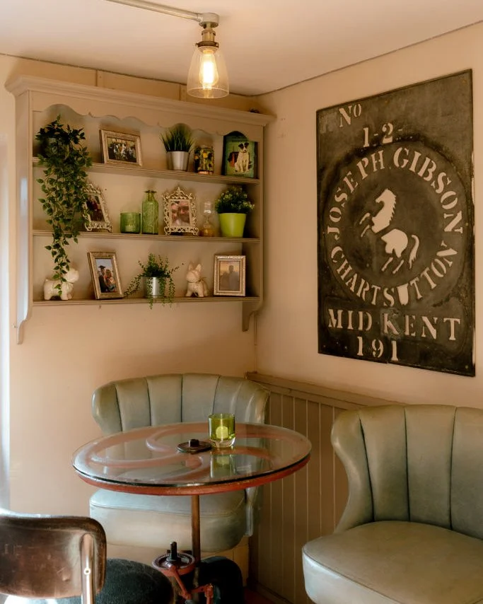 Cozy cafe corner with a glass-topped table, two vintage green upholstered chairs, wall decor including framed photos and plants, and a large black and white poster on the wall.