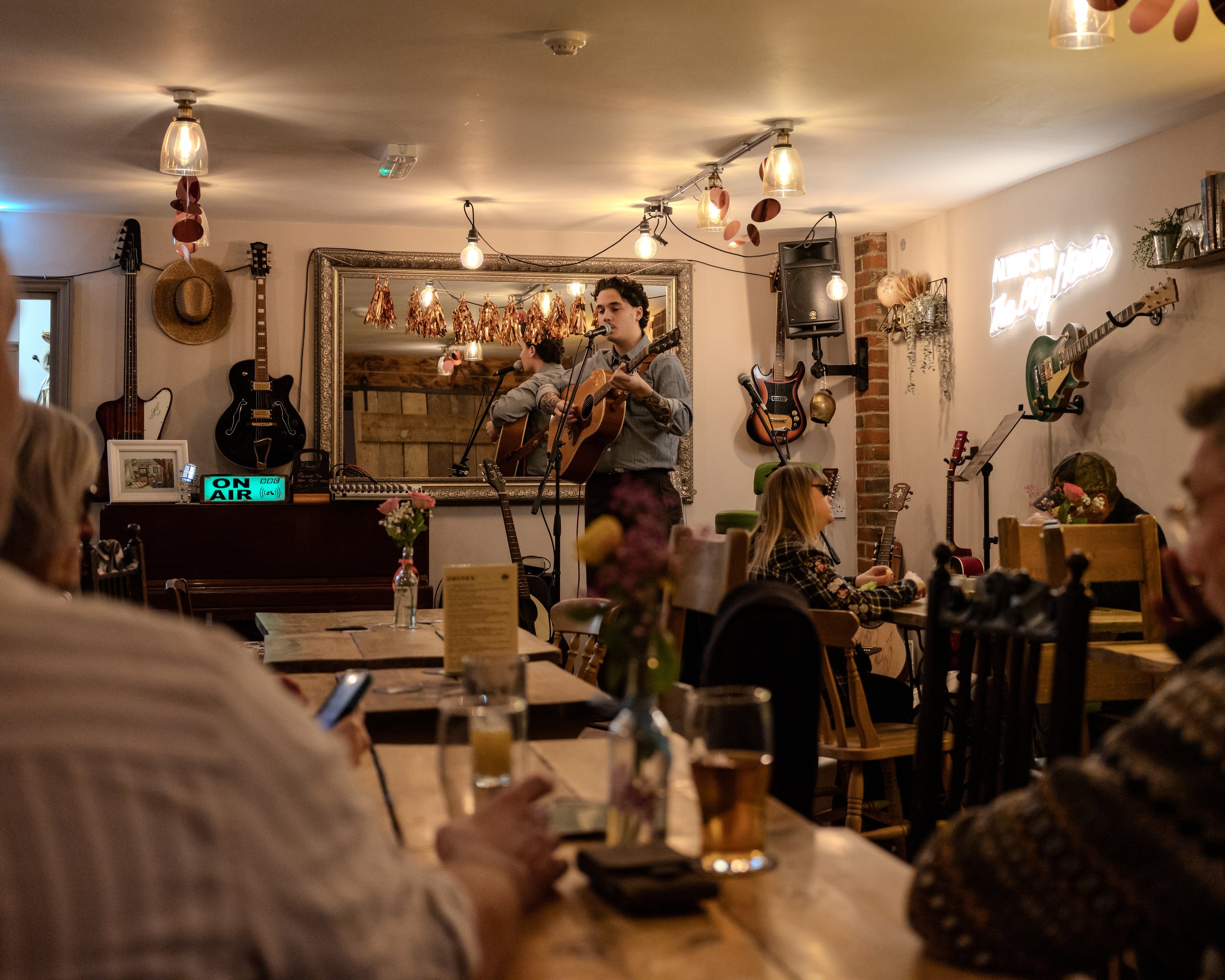 A live music performance in a cozy, decorated indoor venue with a couple of musicians playing guitars and singing, surrounded by an audience at tables with drinks and flowers.