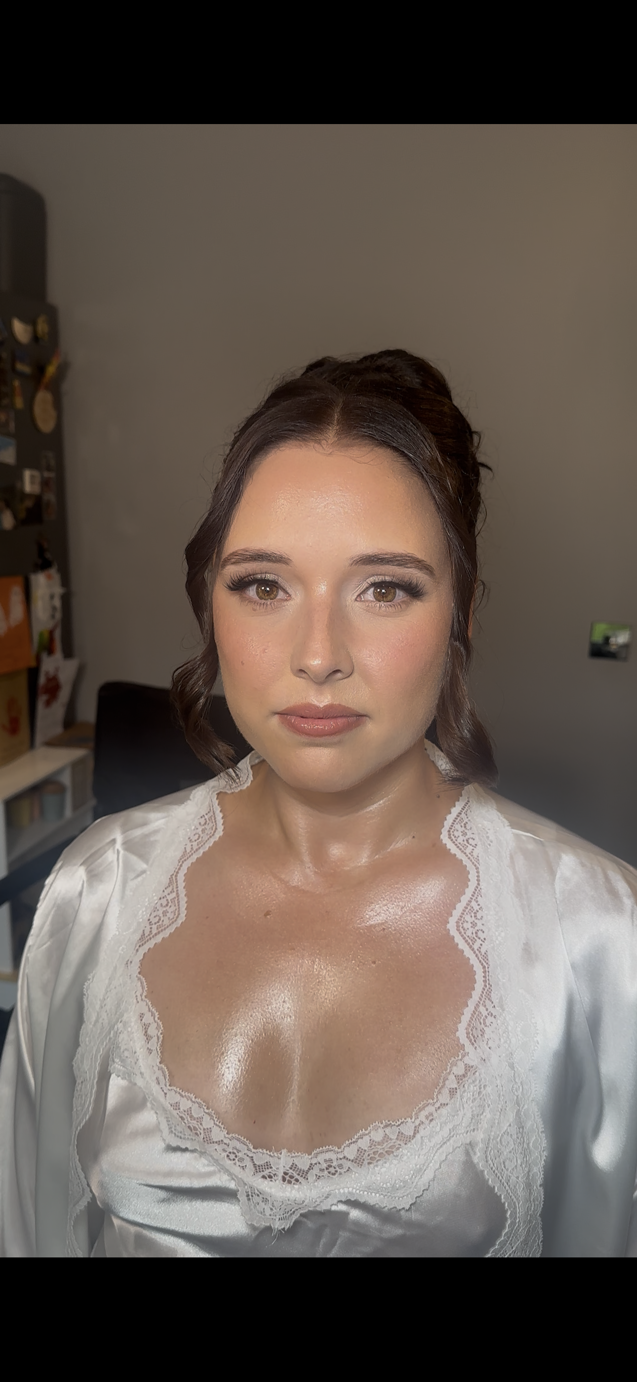 A woman with brown hair styled in loose curls, wearing a white satin and lace garment, and makeup with defined eyebrows and glossy lips. She is indoors, with a neutral background and some household items visible.
