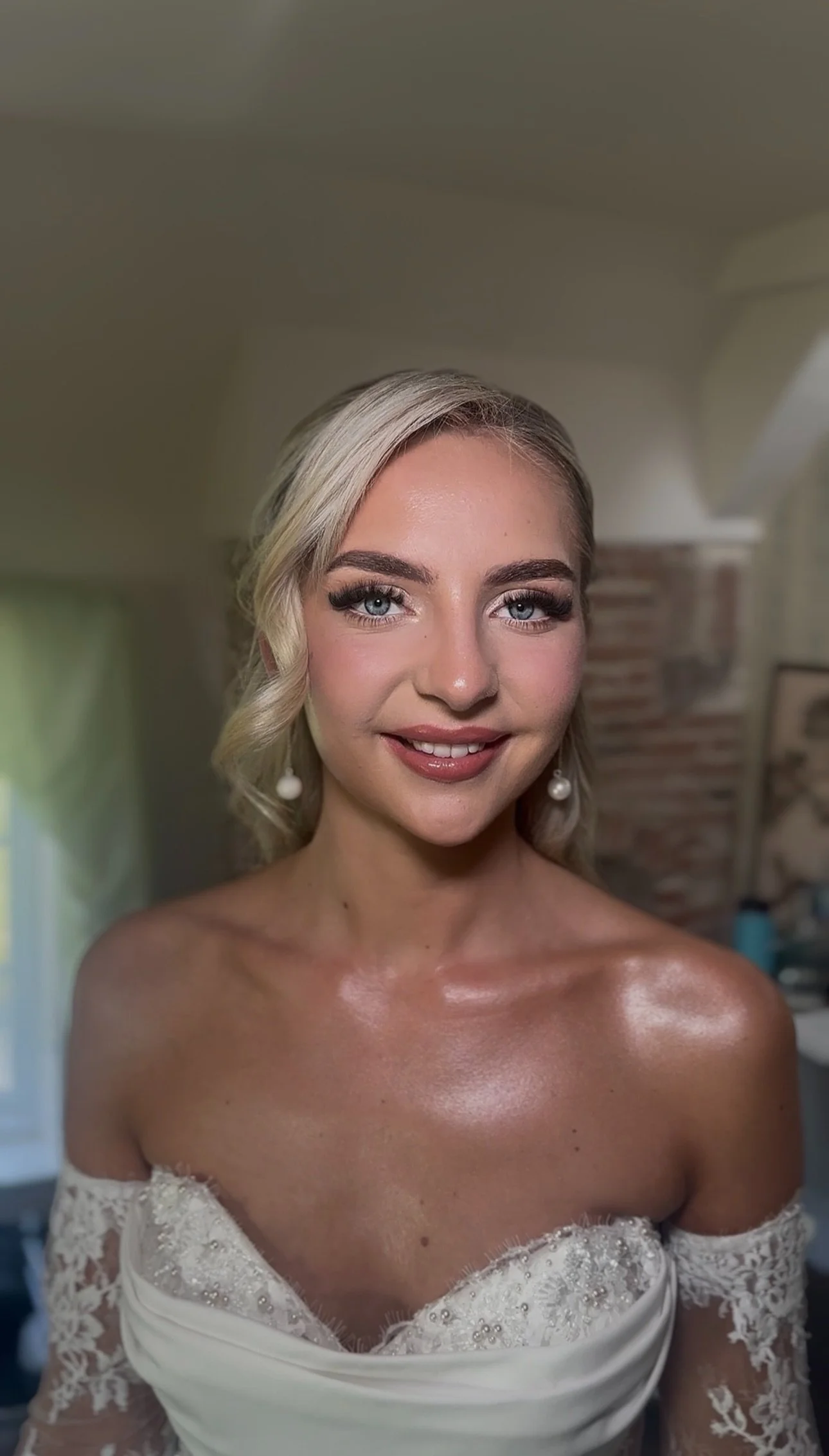 A woman with styled blonde hair, wearing makeup, and pearl earrings, dressed in an off-shoulder white lace dress, smiling in an indoor setting.