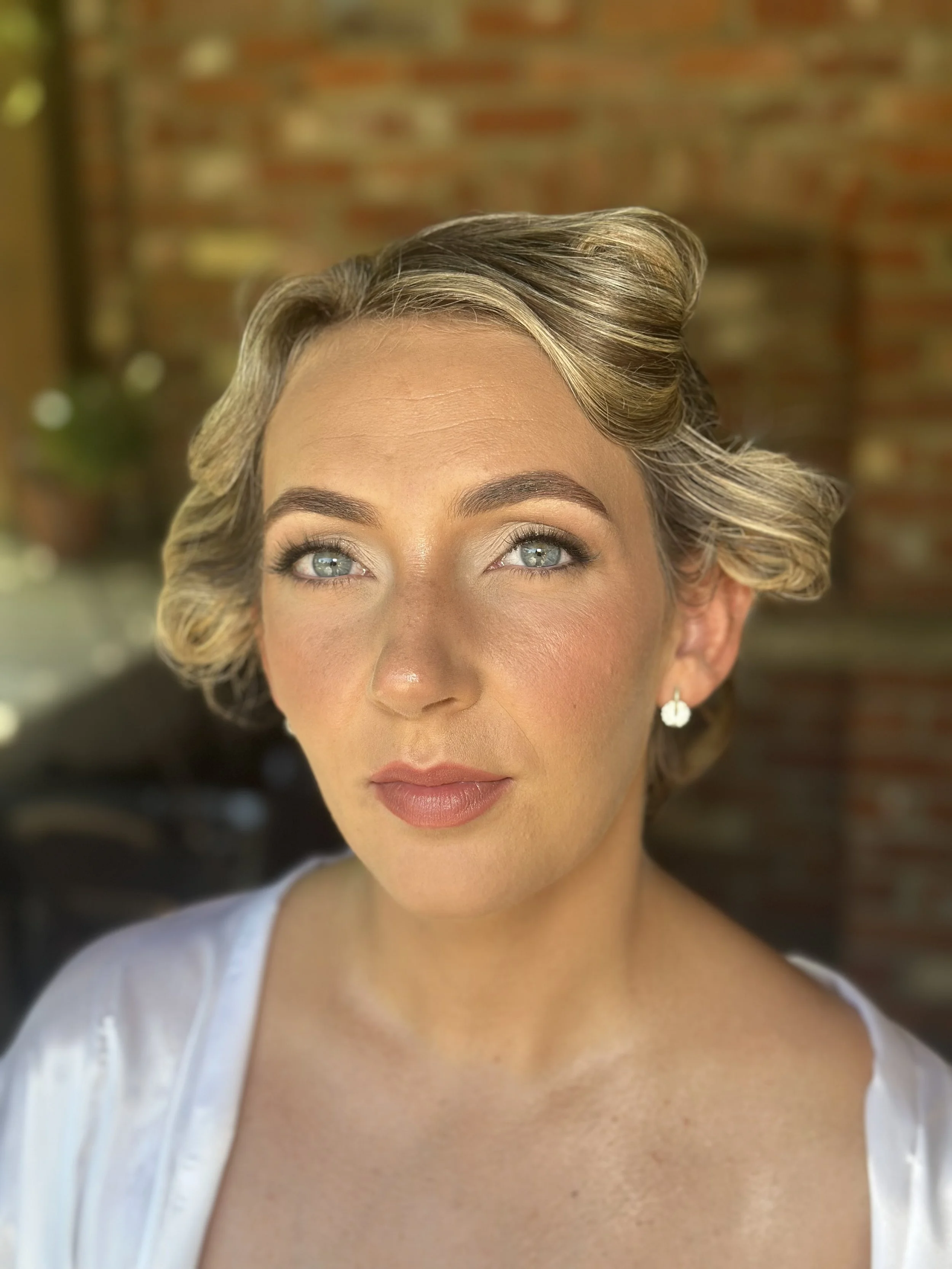 A woman with styled hair, blue eyes, light makeup, and wearing a white top, with a brick wall background.