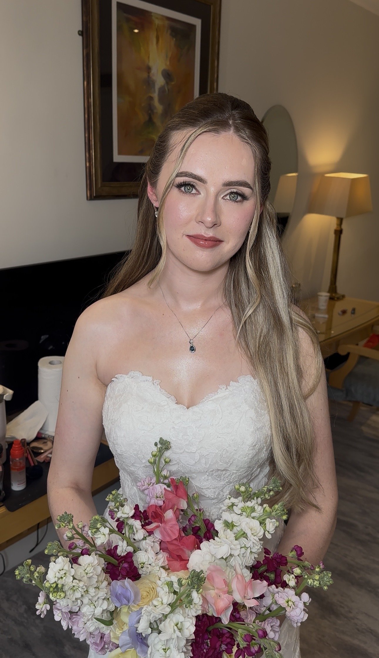 A young woman in a white wedding dress holding a large bouquet of colorful flowers, standing in a room with a painting of a lion and a mirror in the background.