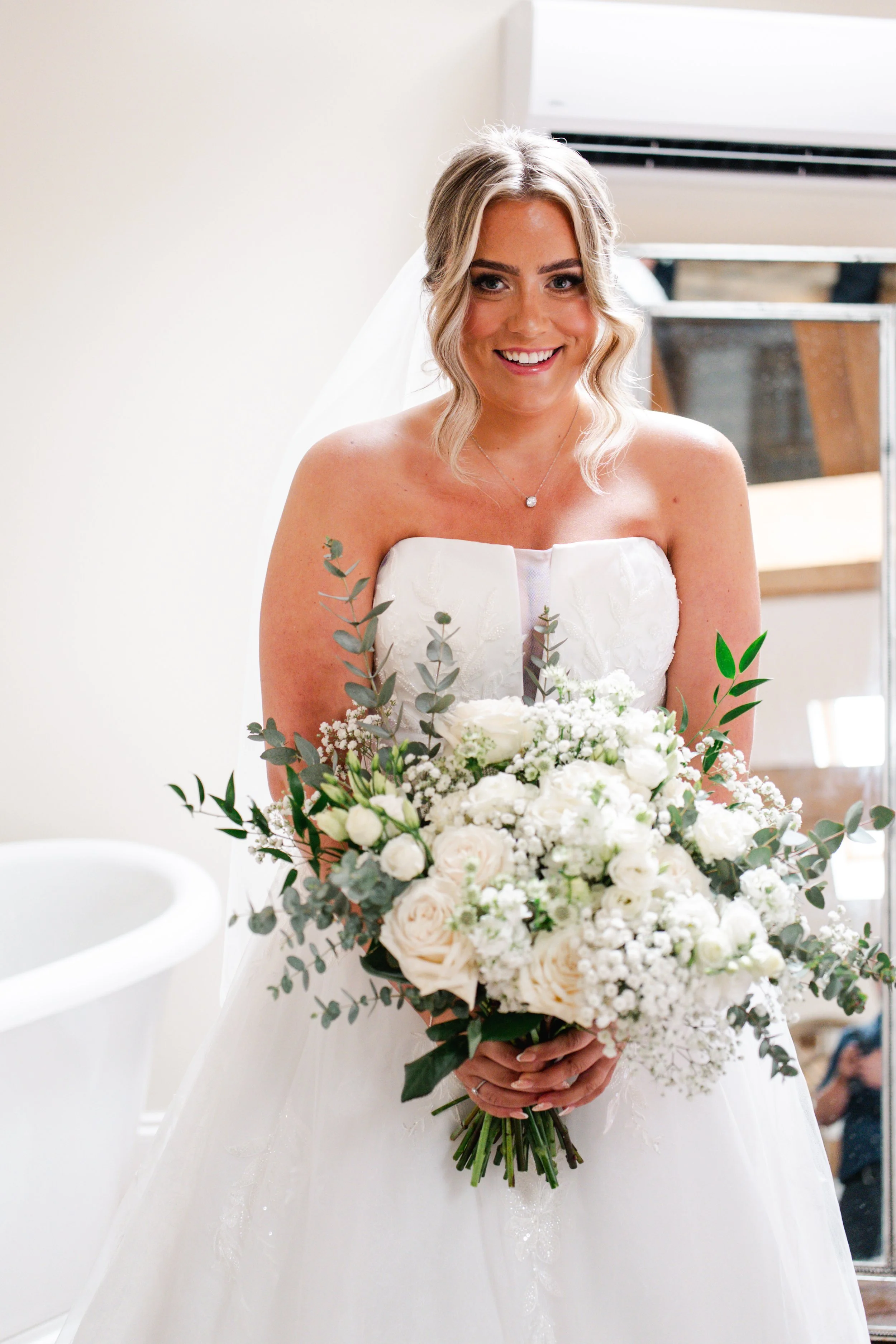 A smiling bride in a strapless white wedding gown holding a large bouquet of white roses, baby's breath, and greenery.
