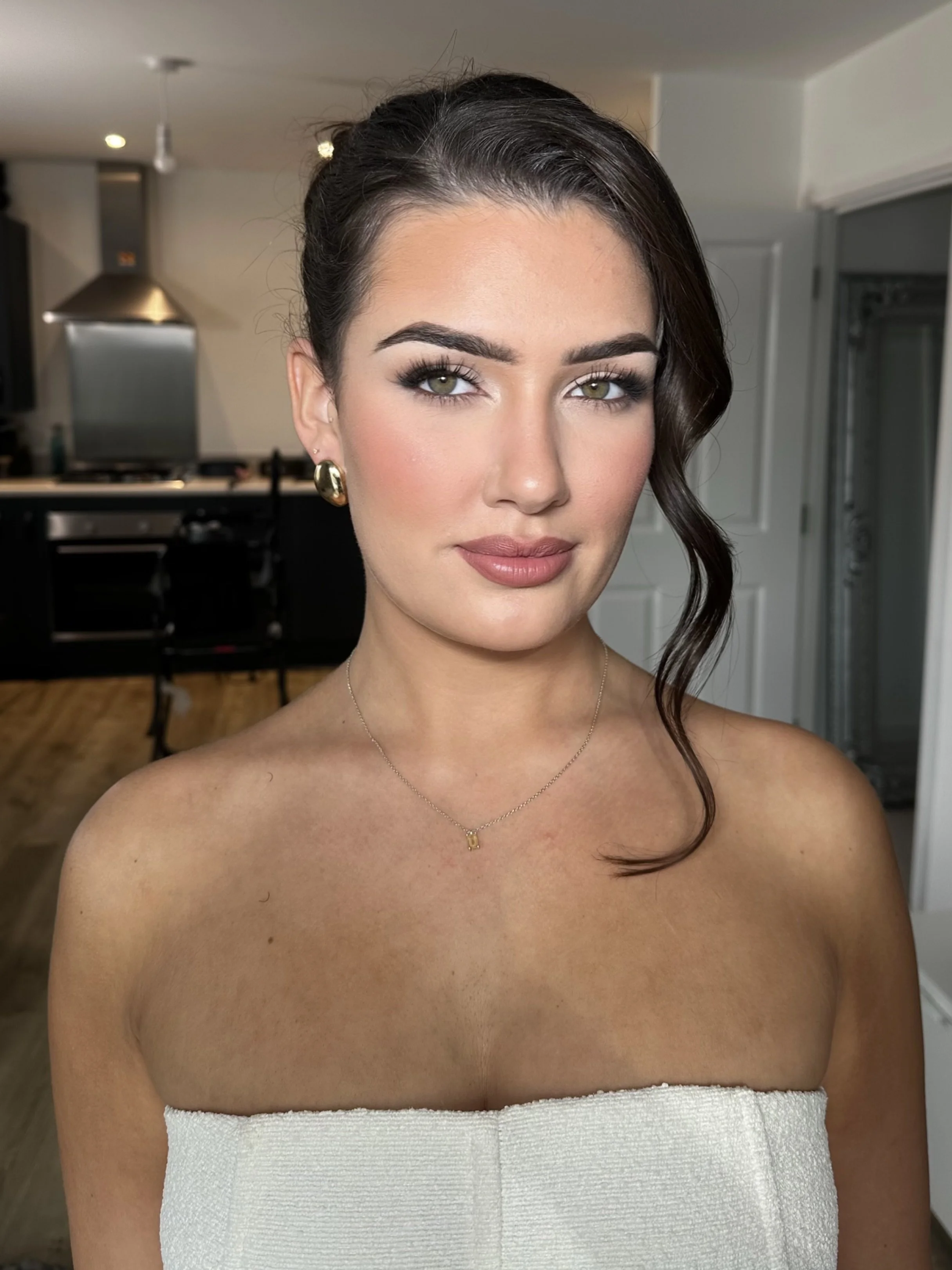 A young woman with dark hair styled in loose curls, wearing gold earrings and a pendant necklace. She has makeup on with defined eyebrows, eye makeup, and nude lipstick. She is wearing a strapless white top. The background shows a modern kitchen with black cabinets and a wooden floor.