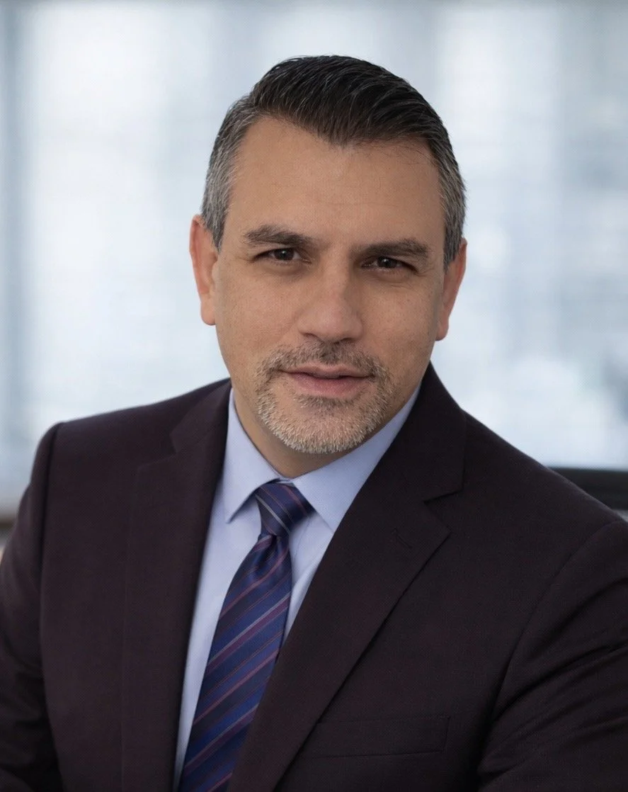 Portrait of a man in a business suit with a light blue shirt and a striped tie, posing in an office environment with blurred windows in the background.