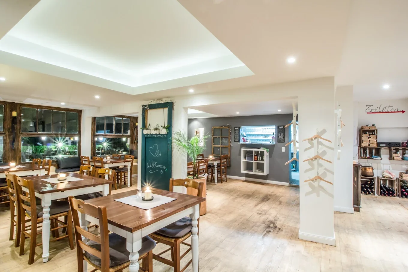 Cozy, well-lit restaurant interior with wooden tables and chairs, decorative plants, and large windows showing outdoor lighting at night.
